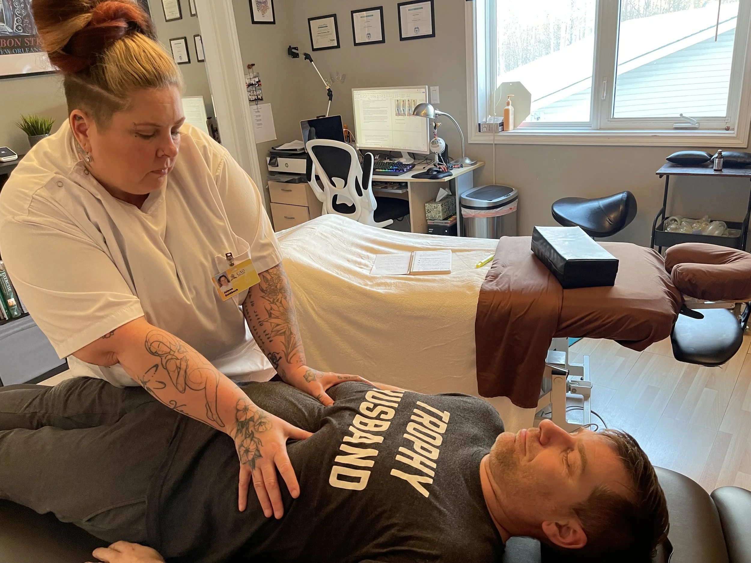 A woman with tattoos on her arms, wearing a beige shirt and a name badge, performs a medical massage or therapy on a man lying face down on a treatment table. The man is wearing a black t-shirt with white text. The room appears to be an office or treatment room with a computer, framed certificates, and a window providing natural light.