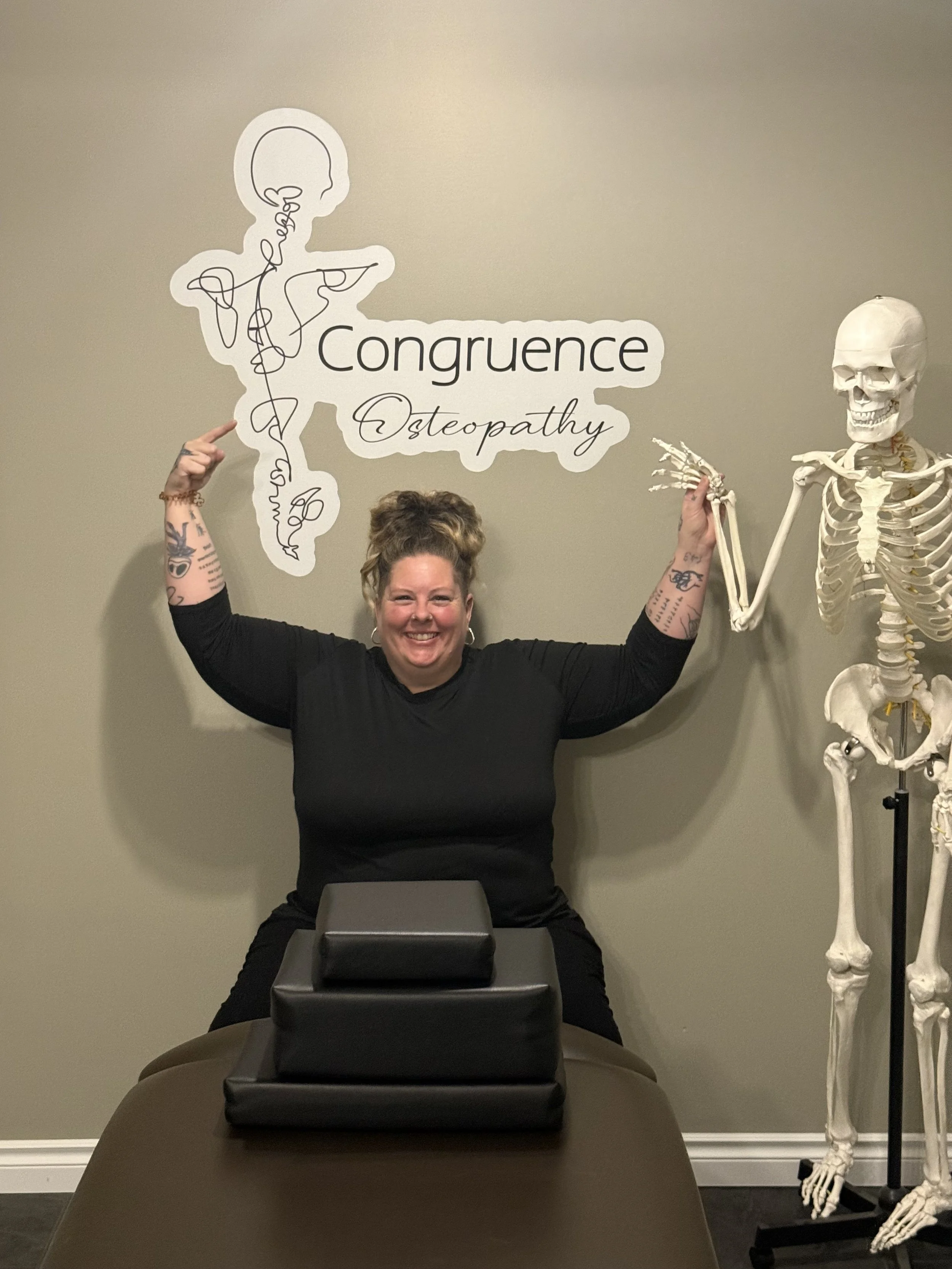 A woman with tattoos on her arms smiling and pointing to a sign that reads 'Congruence Osteopathy' and a human skeleton model standing next to her in an office or clinic setting.