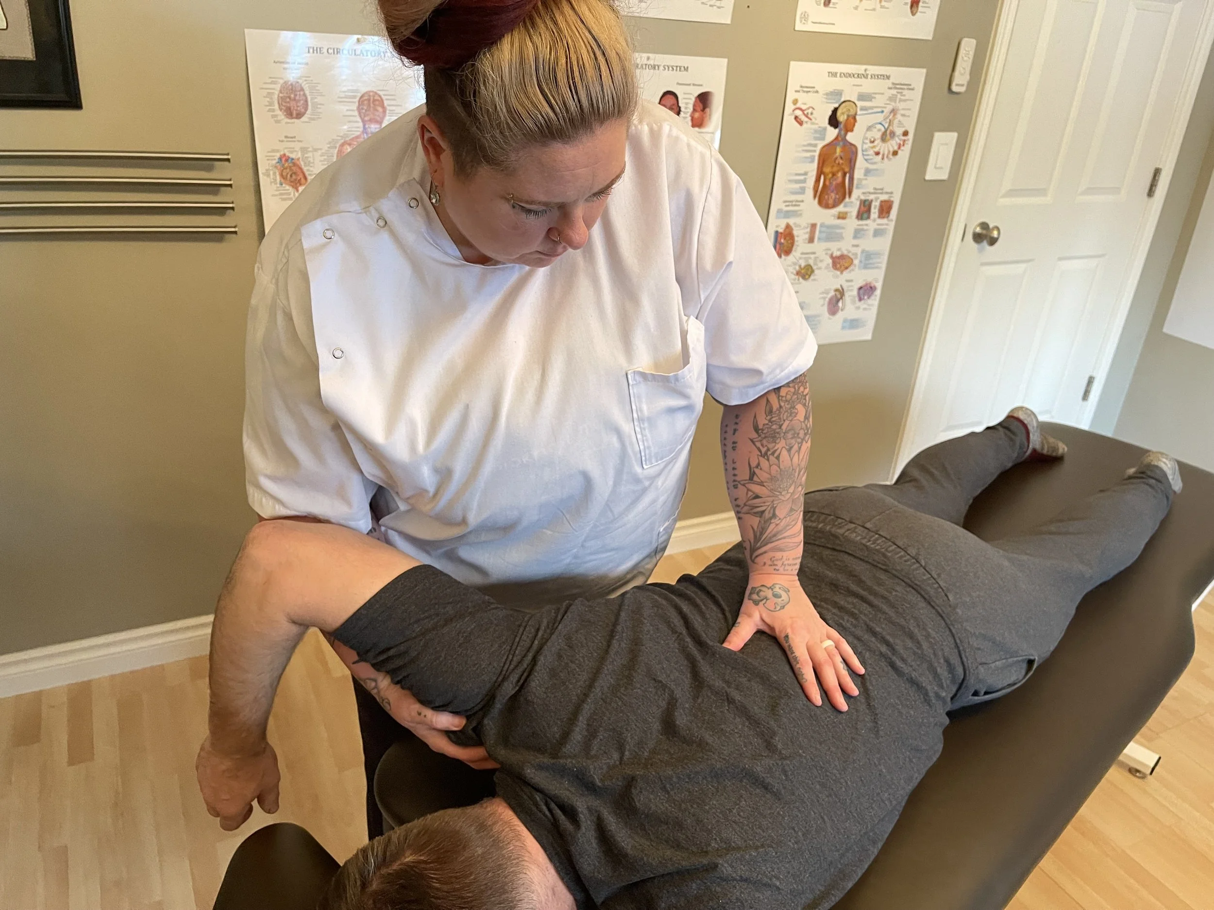 A person in white medical scrubs performs a chiropractic adjustment on a person lying face down on a massage table. The scene is in a room with medical charts on the wall.