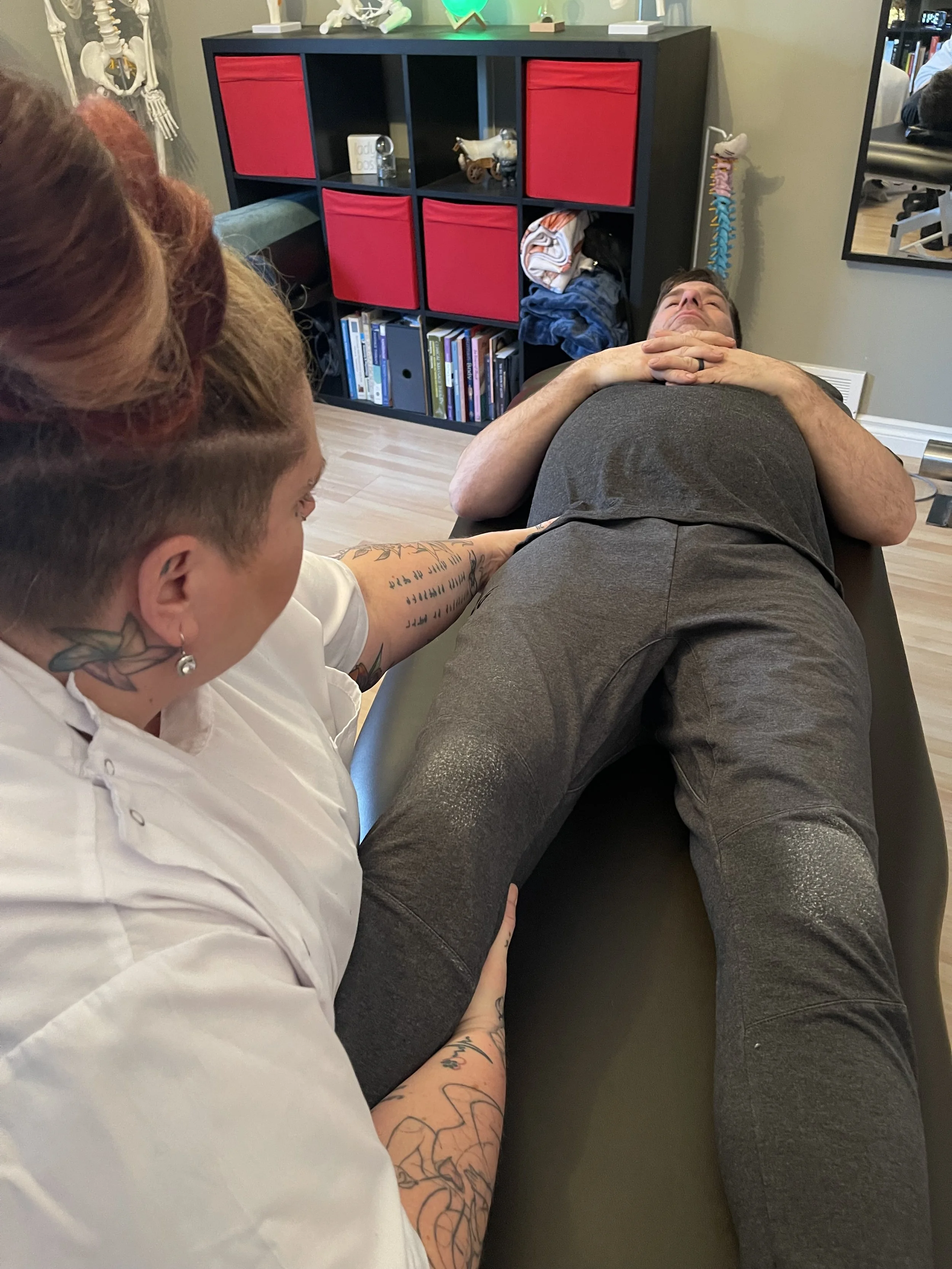 A person lying on a chiropractic table while a healthcare professional examines their lower back. The setting appears to be a medical or chiropractic clinic with a bookshelf, toys, and anatomical models in the background.