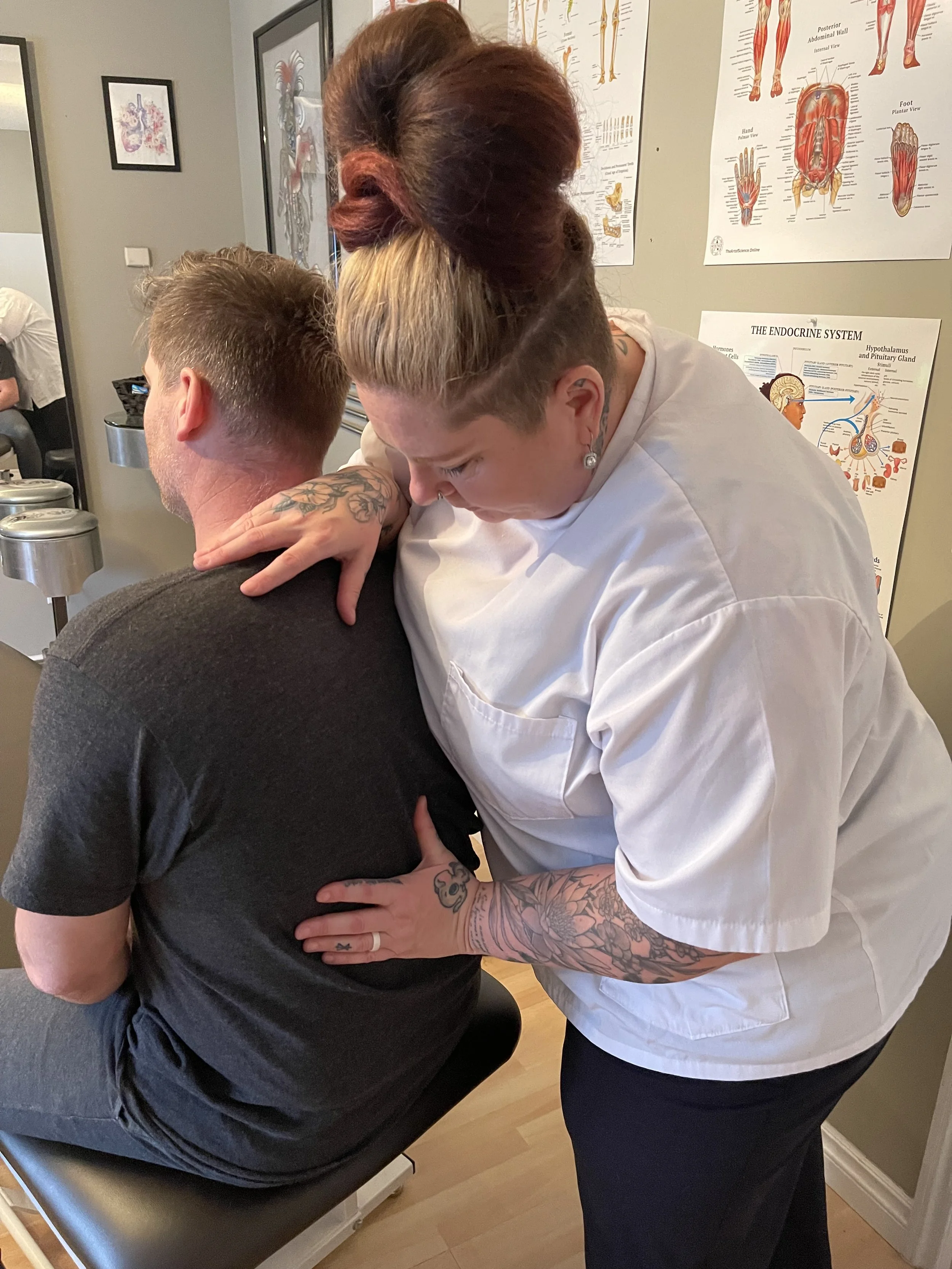 A woman with tattoos on her arms, wearing a white medical uniform, is giving a shoulder massage to a man with short brown hair, who is seated on a table. The scene takes place in a room with anatomy posters on the wall.