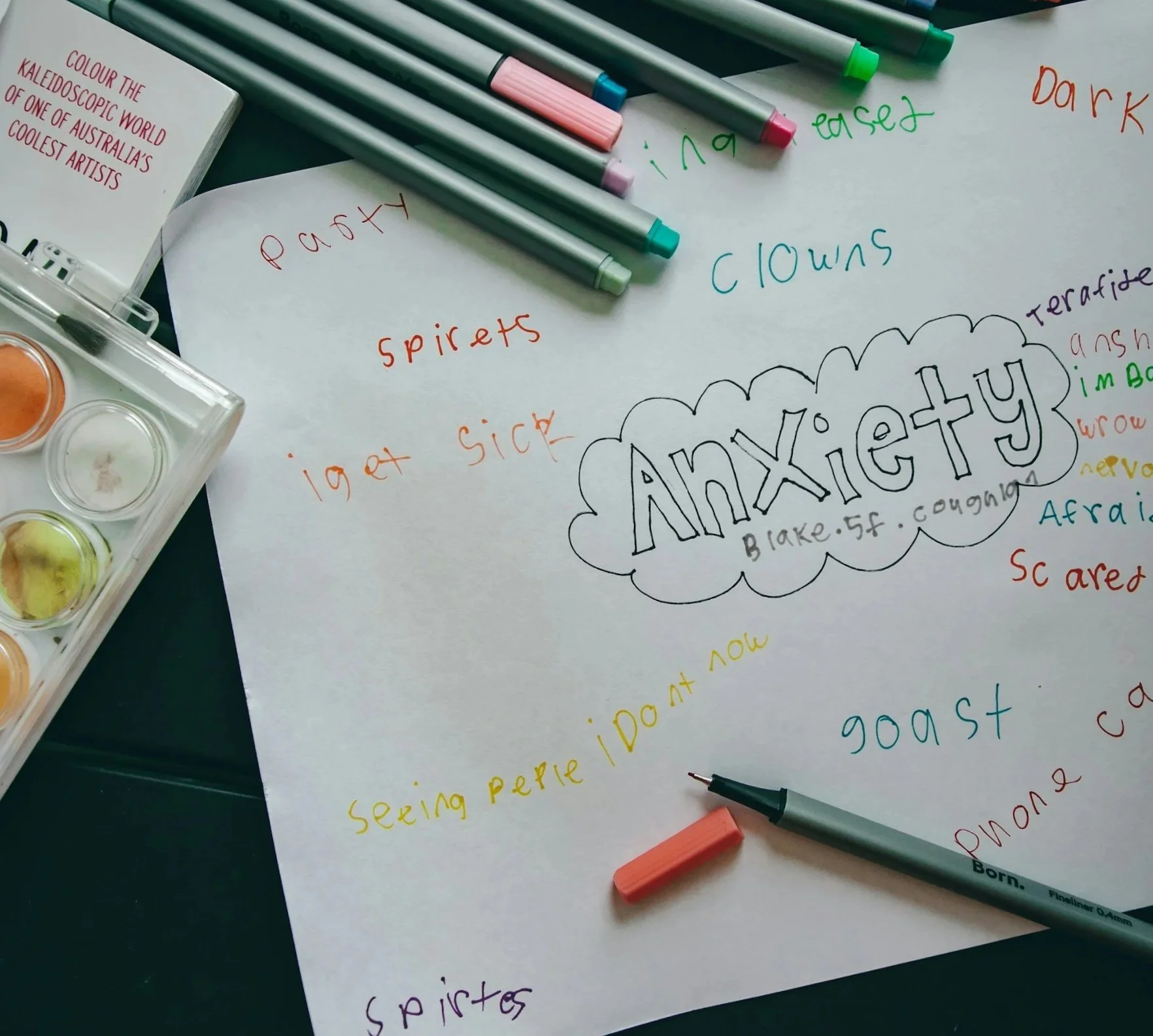 A sheet of white paper with handwritten colorful words, surrounded by markers and a watercolor palette. The words include "colour the kaleidoscopic world of one of Australia's coolest artists," "anxiety," "spirits," "light sick," and "anthiexiety." The words are in various colors including red, green, blue, orange, and purple, with some words underlined or styled creatively.
