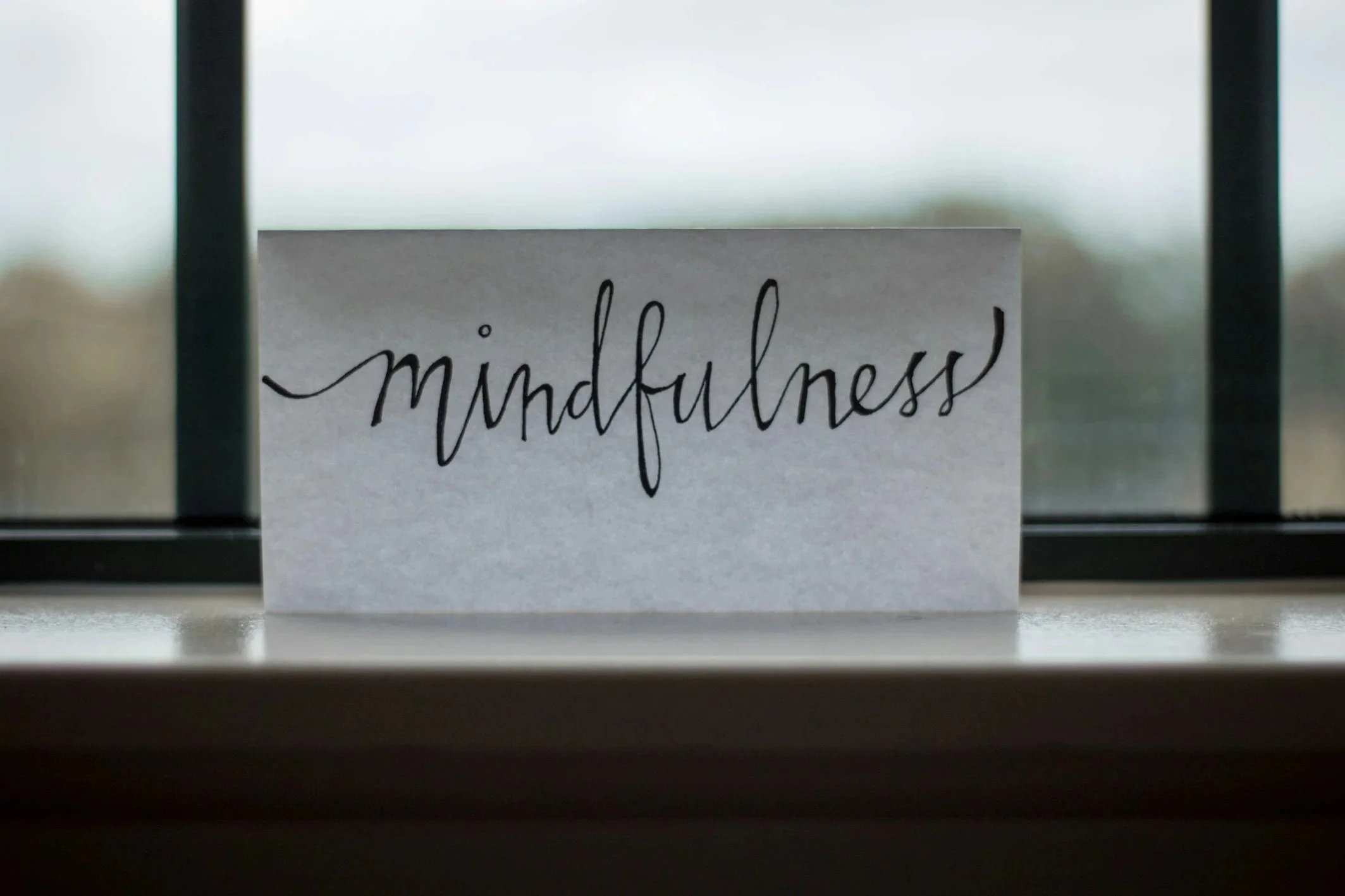 A piece of paper with the word 'mindfulness' written in black cursive is placed on a surface, viewed through a glass window or mirror.