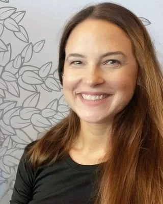 A woman with brown hair smiling, standing in front of a wall with a leaf pattern.