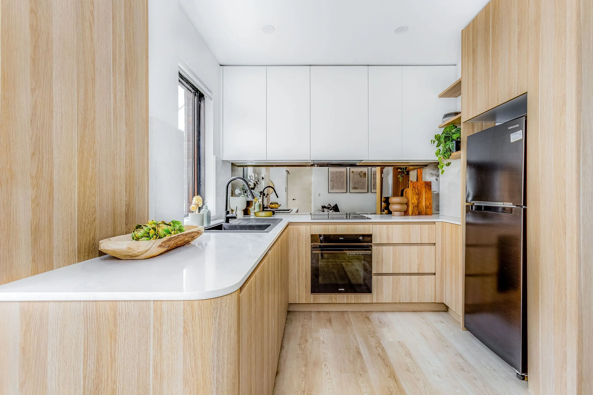 Modern kitchen with natural wood cabinetry, white countertops, black appliances, and a window with natural light.