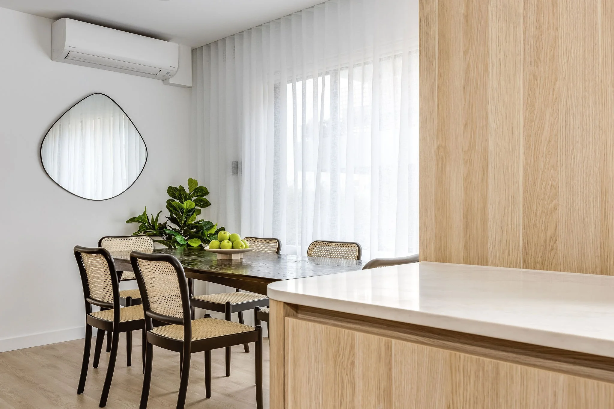 Dining room with a wooden table, six chairs, a bowl of green apples, a potted plant, a white wall with an abstract mirror, sheer white curtains, and an air conditioning unit.