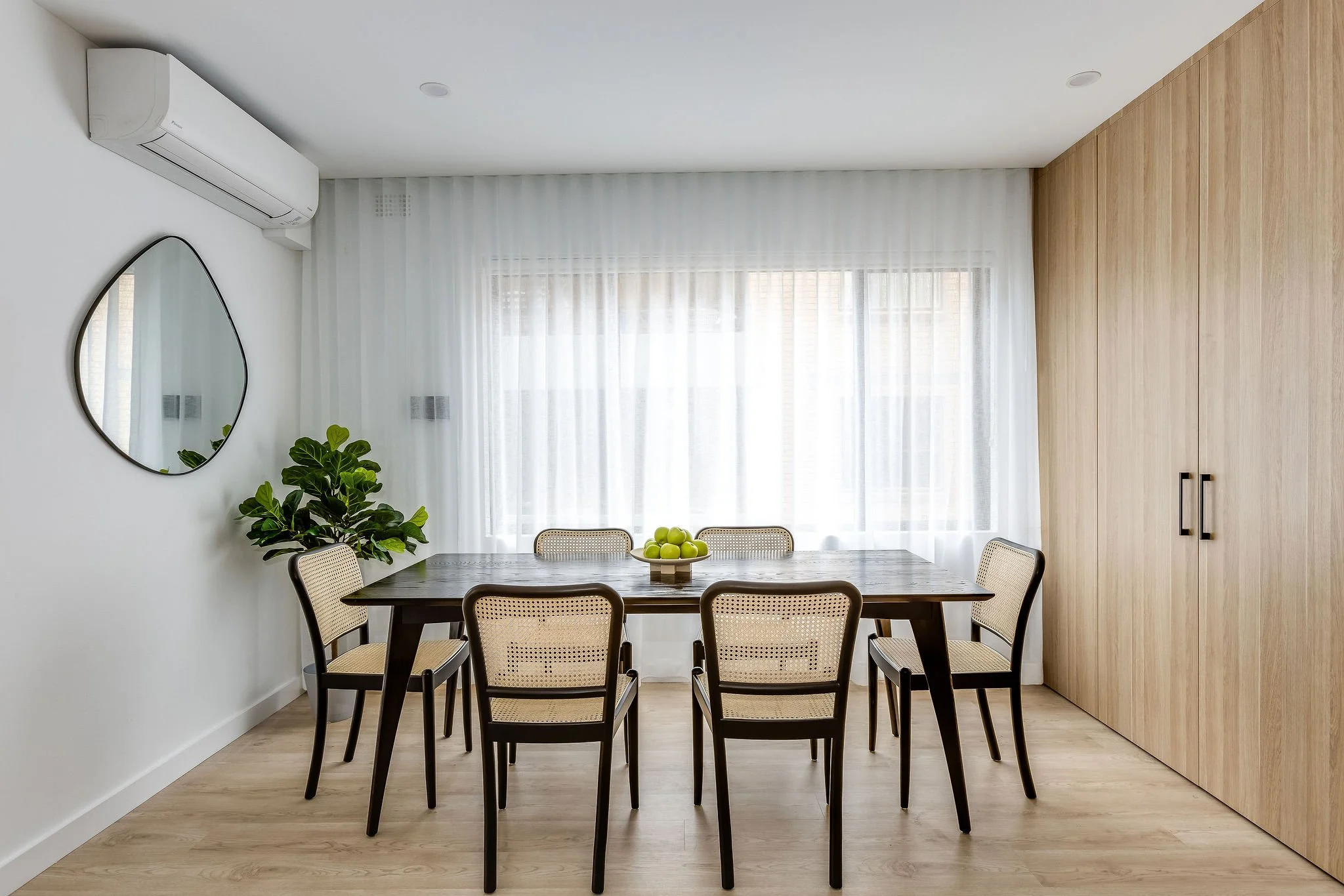 A modern dining room with six chairs around a dark wood table, a bowl of green apples in the center, a large window with sheer white curtains, a mirror on the white wall, a potted plant, an air conditioning unit, and a wooden closet door.