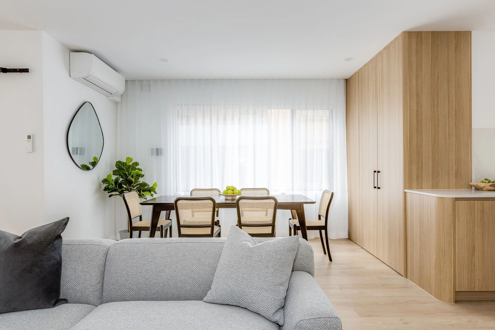 Modern living room with a gray sofa, white cushions, a wooden dining table with six chairs, a large window with sheer curtains, a wall-mounted mirror, a potted plant, and wooden cabinets.