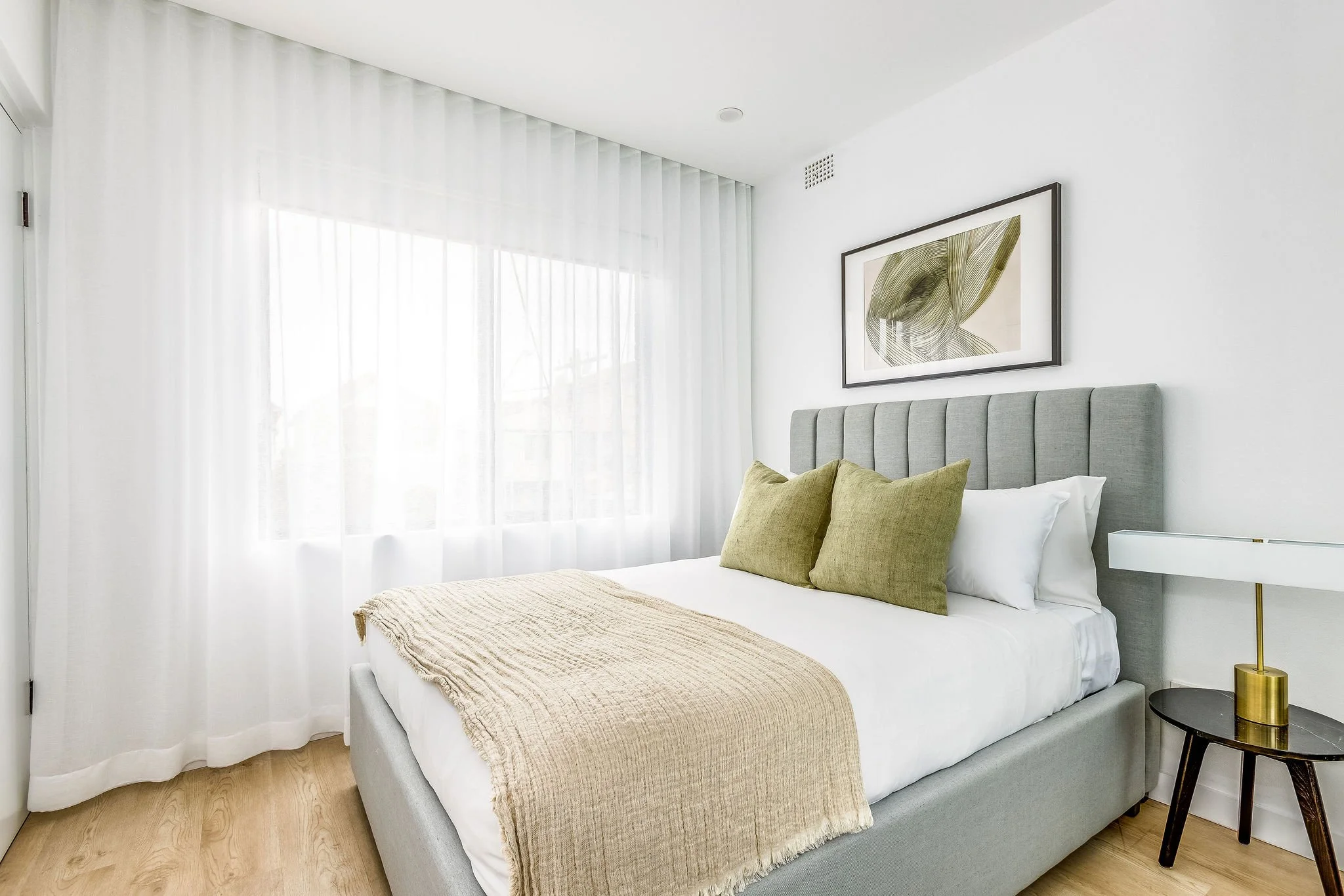 A neatly made bed with a gray upholstered headboard, white bedding, two green pillows, and a beige throw blanket in a bright, modern bedroom with white walls, sheer white curtains, a framed botanical print, and a small black bedside table with a gold lamp.
