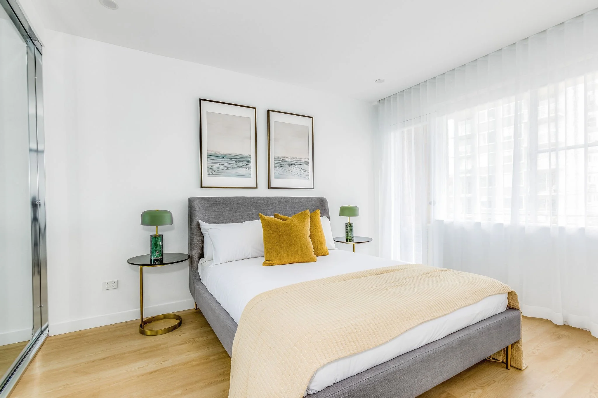 Modern bedroom with a gray upholstered bed, white bedding, and yellow pillows. Two green bedside lamps on black and gold tables, light wooden floors, and white walls with two landscape paintings above the bed. Large window with sheer white curtains.