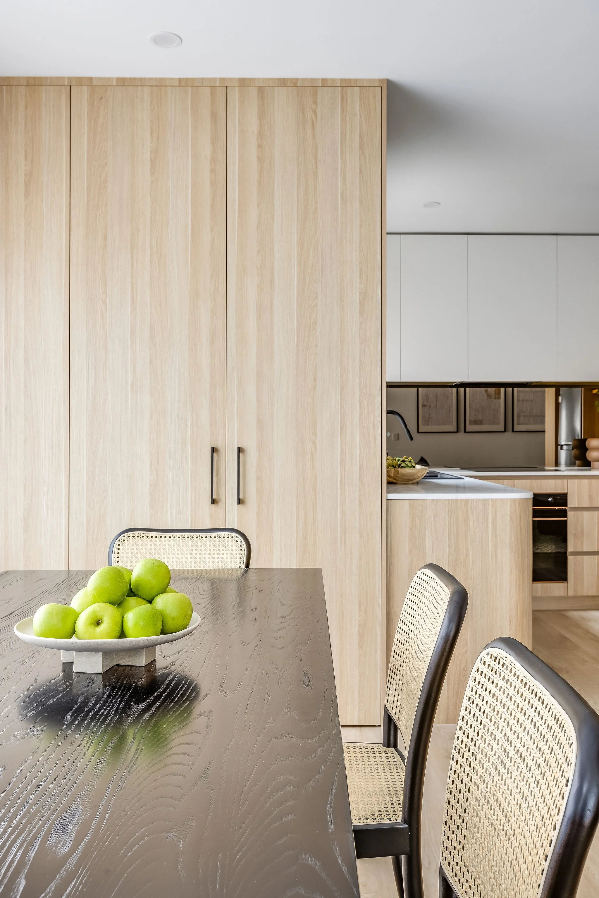 A modern kitchen and dining area with a dark wood table, a bowl of green apples, and light wood cabinetry in the background.