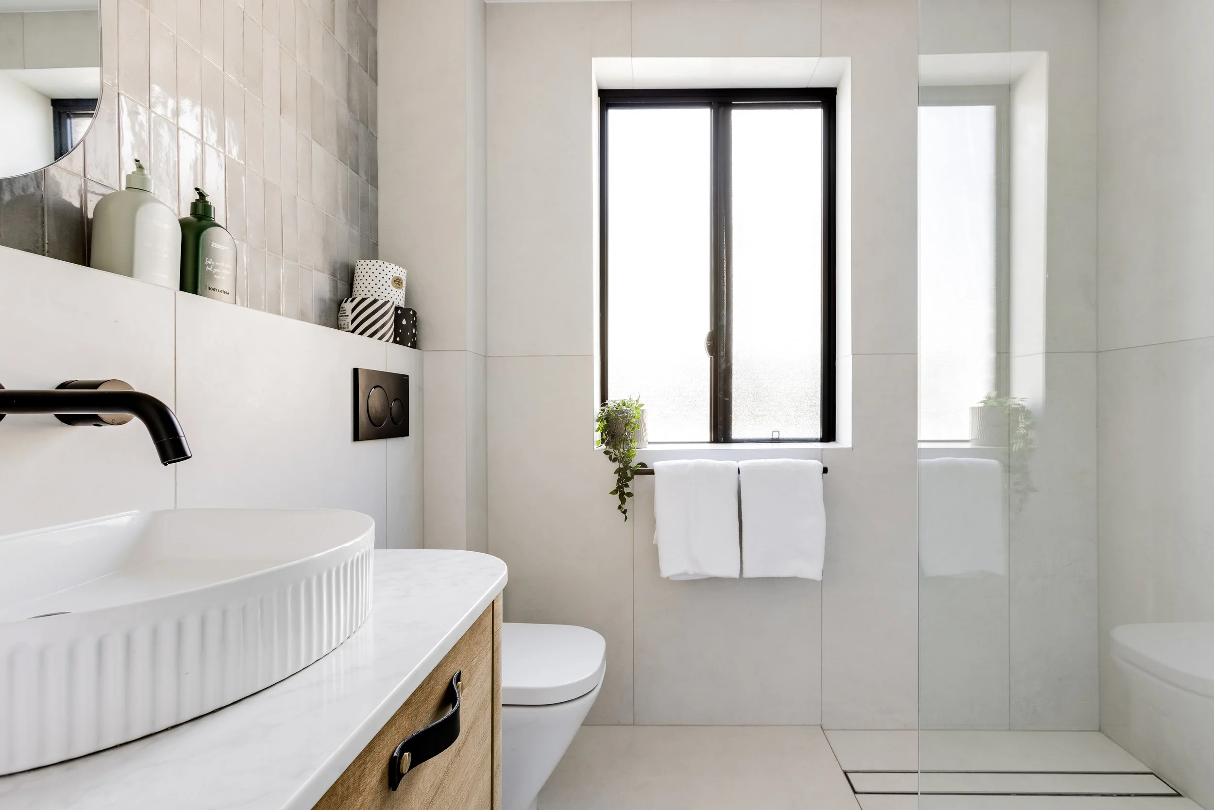 Minimalist bathroom with white walls, black-framed window, white towels, wooden vanity with sink, black faucet, and shower area with glass door.