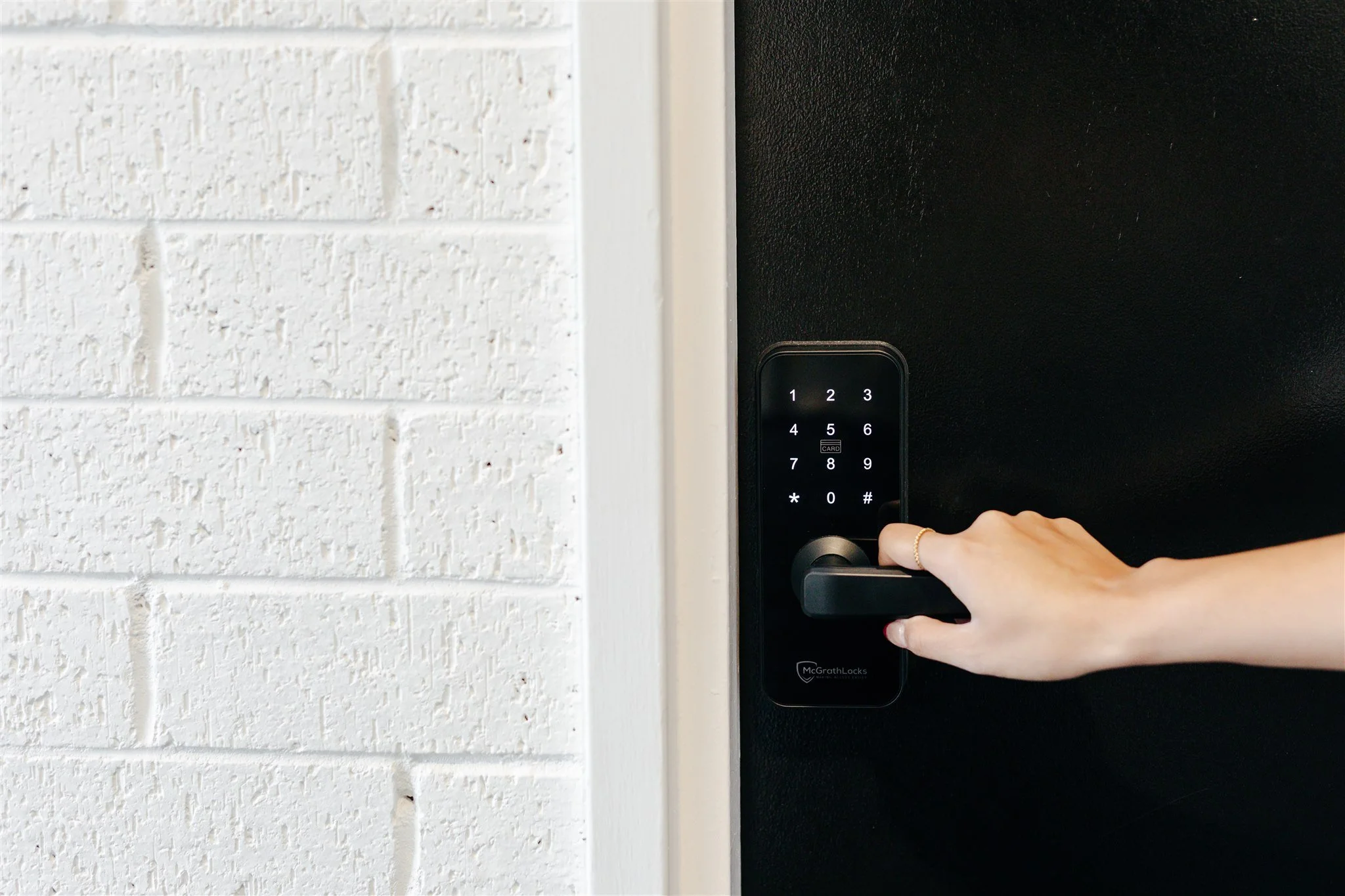 A hand pressing a keyless entry door lock on a black door next to a white brick wall.