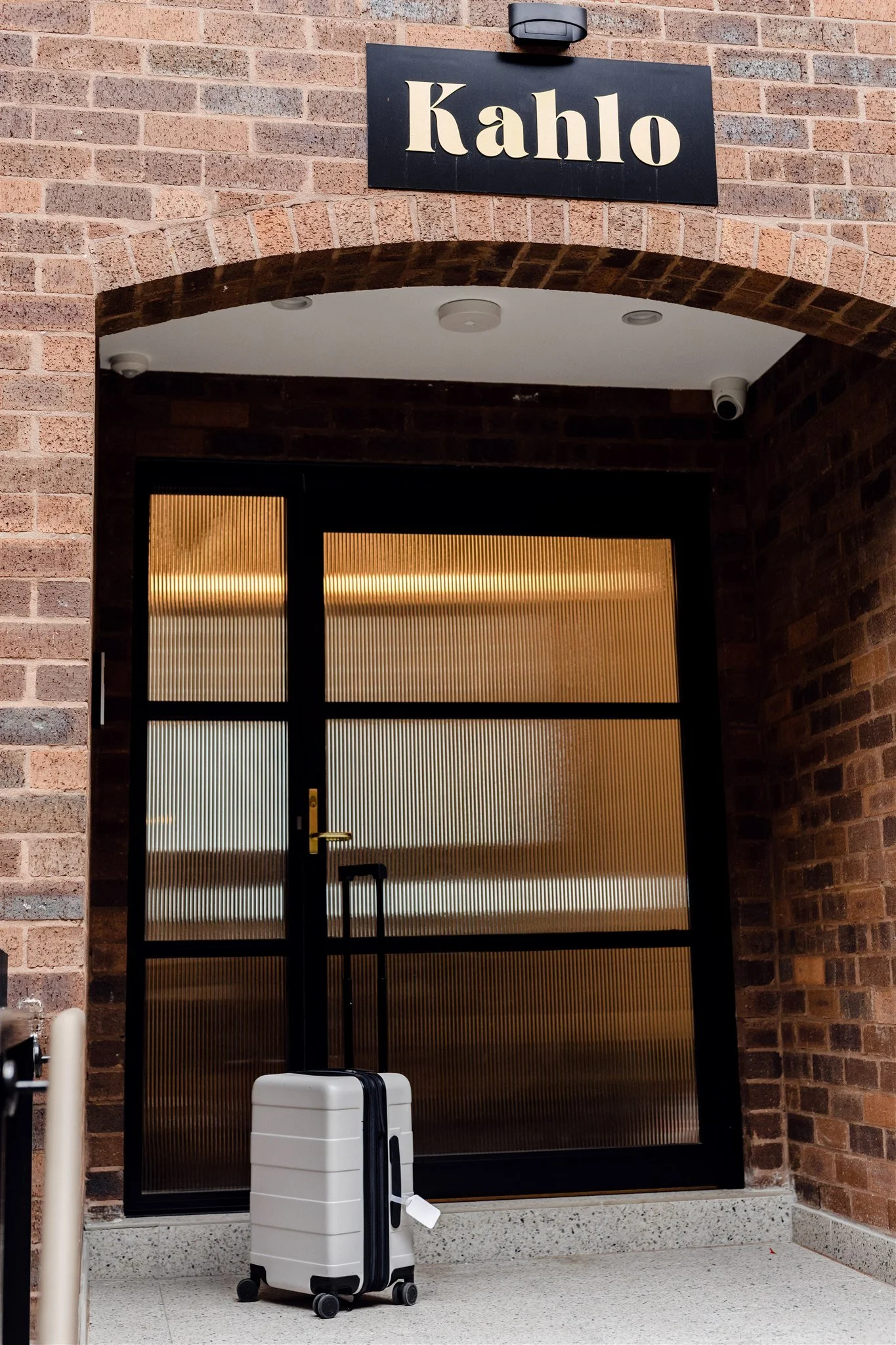 A black sign with white lettering that reads 'Kahlo' hangs above an entryway with a glass door and a brick arch. A white suitcase with a black handle and a black strap is placed in front of the door.