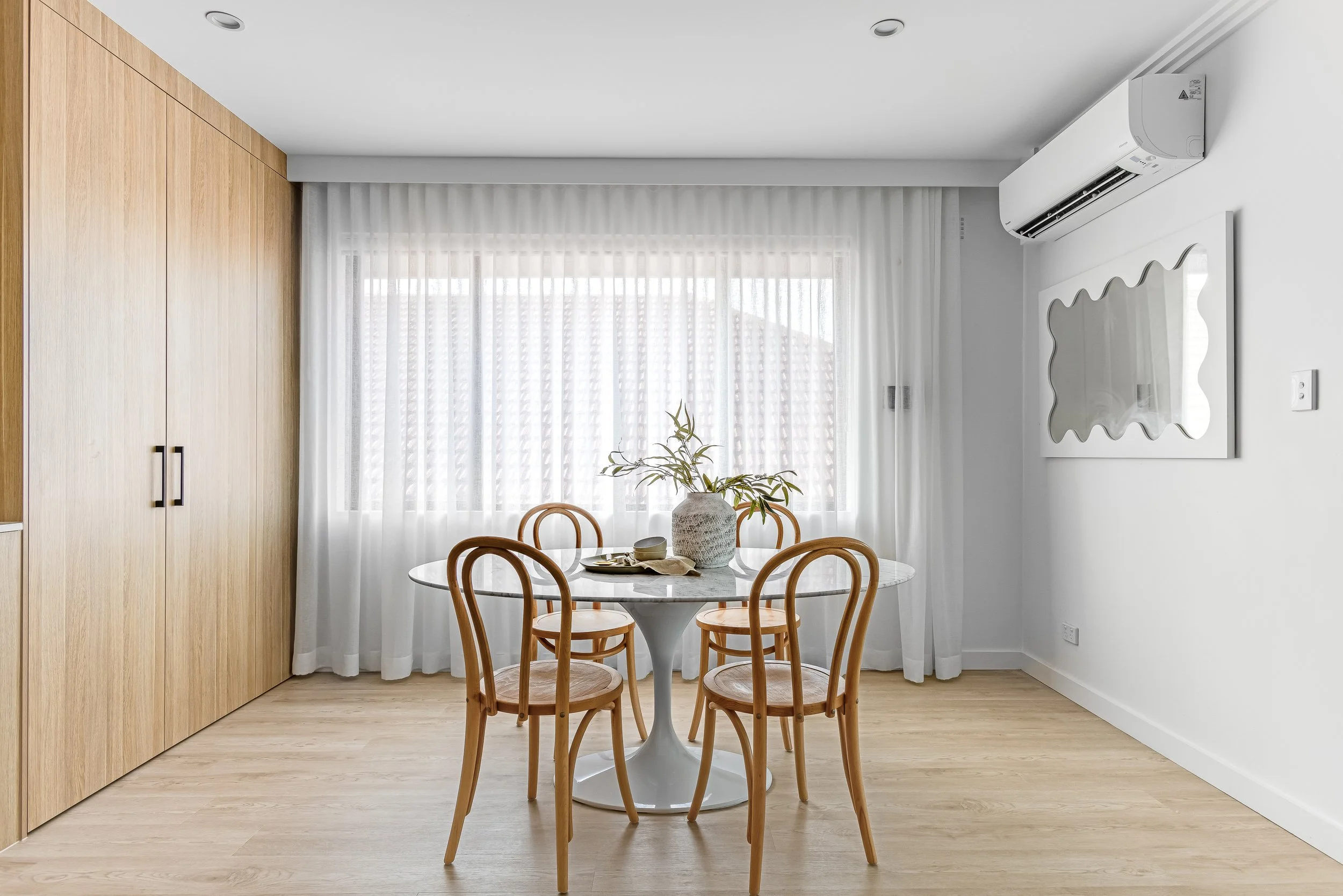 A minimalist dining area with a round white table, four wooden chairs, a white vase with plants, sheer white curtains, and an air conditioning unit on a white wall.
