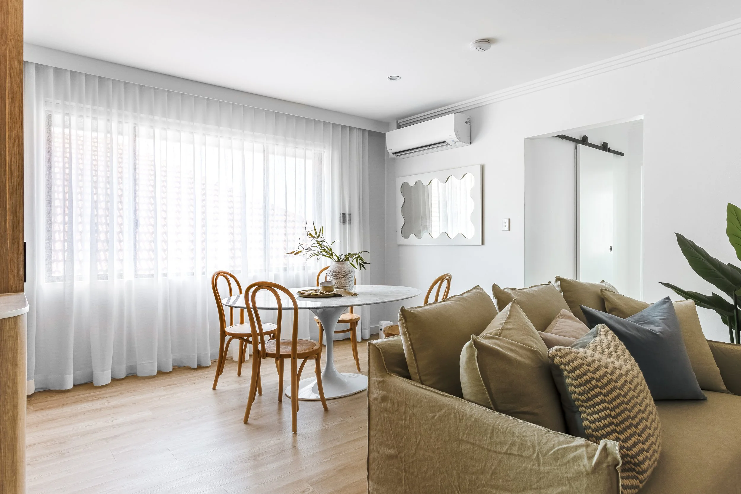 Living room with beige sofa, decorative pillows, wooden dining table with four chairs, potted plant, large window with sheer white curtains, wall mirror, air conditioning unit, and sliding door.