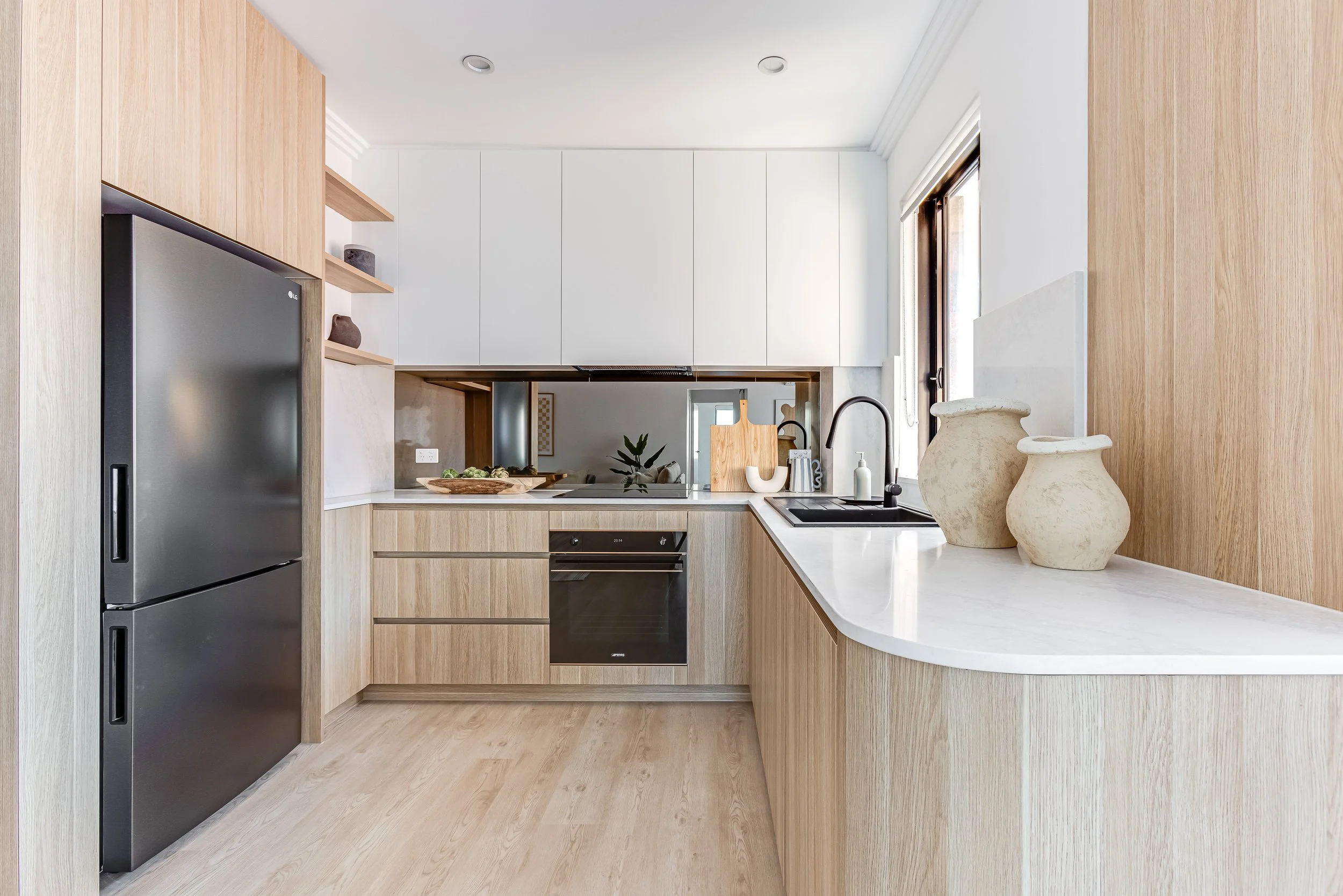 Modern kitchen with light wood cabinetry, white countertops, black sink and faucet, stainless steel oven and refrigerator, and decor vases on the counter near a window.