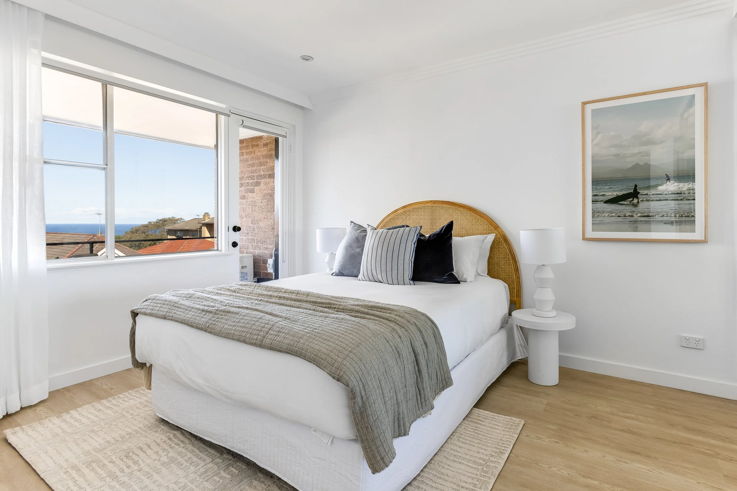 A bright bedroom with a bed, pillows, and a nightstand next to a window showing ocean views.