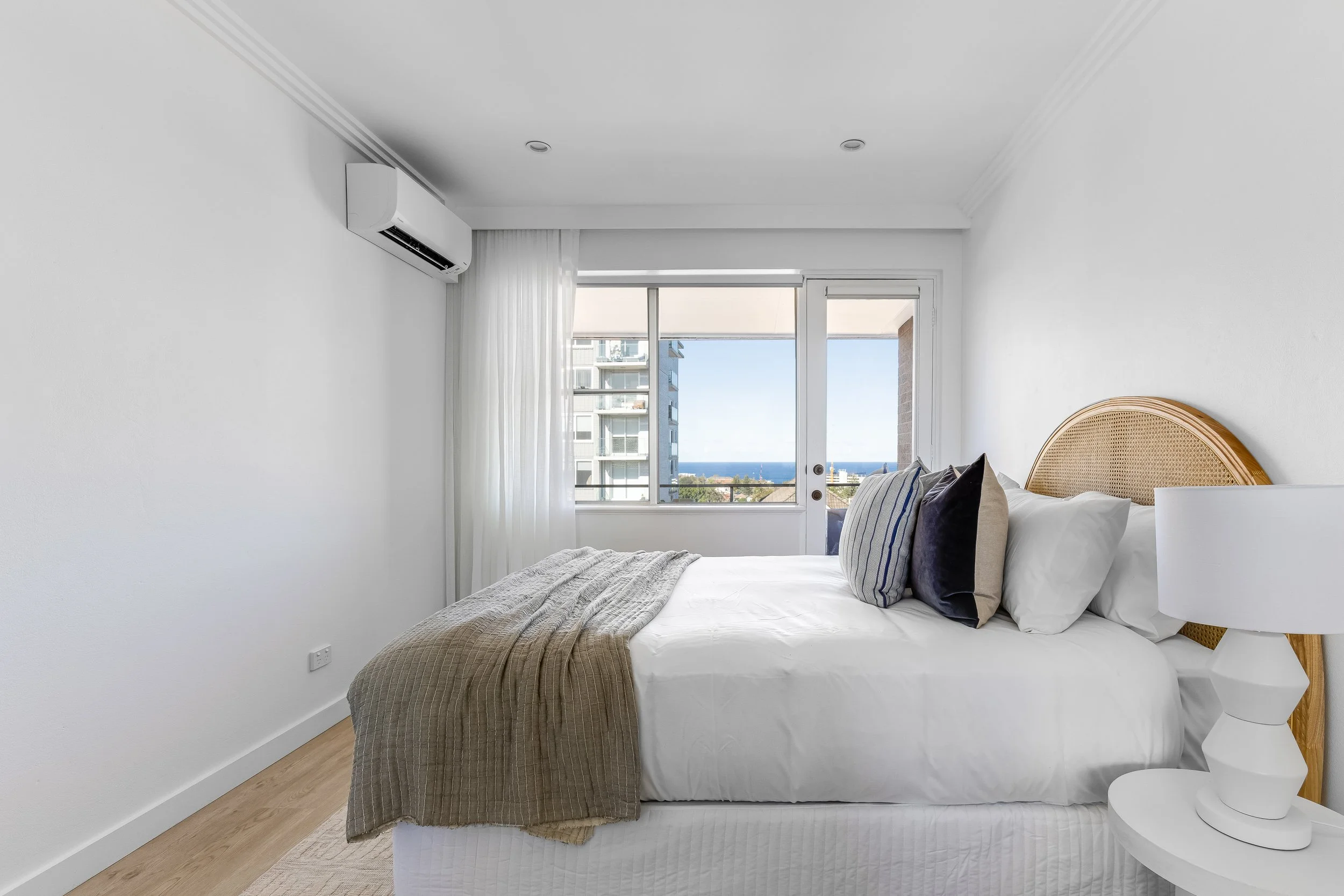 Minimalist bedroom with white walls, a bed with white linens and multiple pillows, a wicker headboard, a white bedside table, a modern white lamp, and a large window with sheer white curtains showing a cityscape and ocean view.