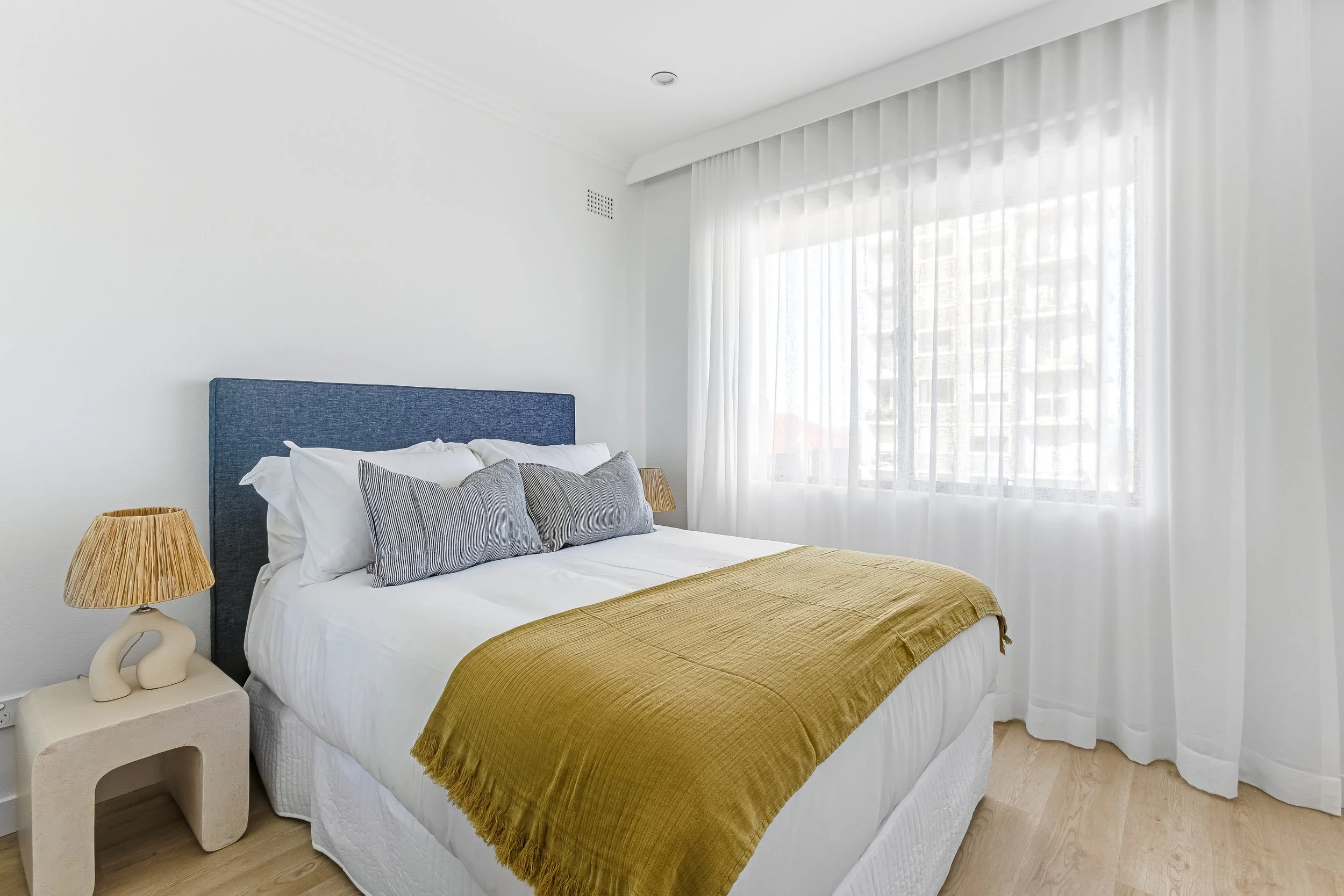 A neatly made bed with white bedding and gray pillows, a blue headboard, and a yellow throw blanket. A bedside table with a unique cream-colored lamp is beside the bed. Large window with white curtains allows natural light into the room.