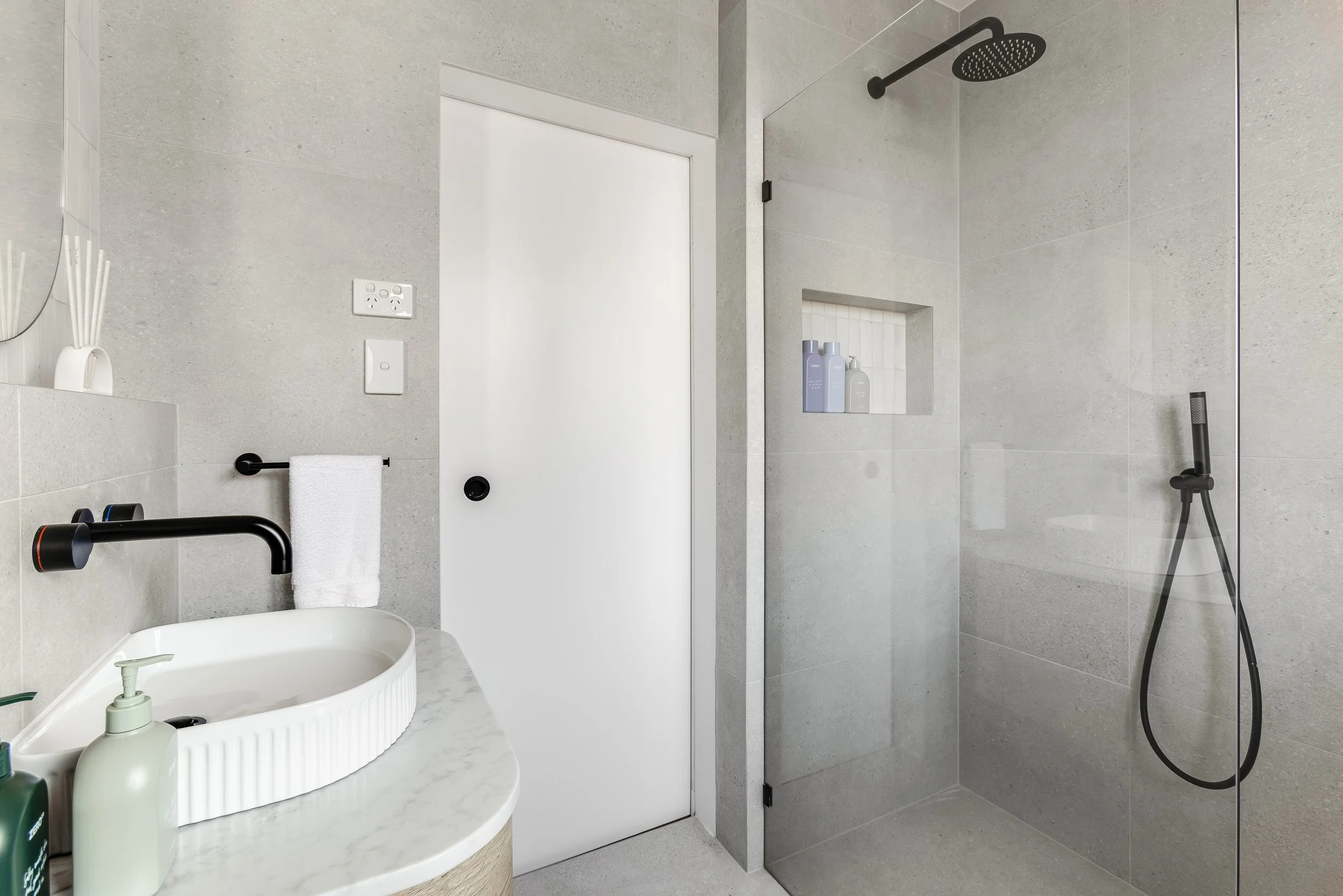 Modern bathroom with a shower area featuring a rain shower head and a daftar shower hand held attachment, a small niche in the wall with toiletries, a gray countertop with a vessel sink, black wall-mounted faucet, and bathroom essentials, a white door, and gray tiled walls.