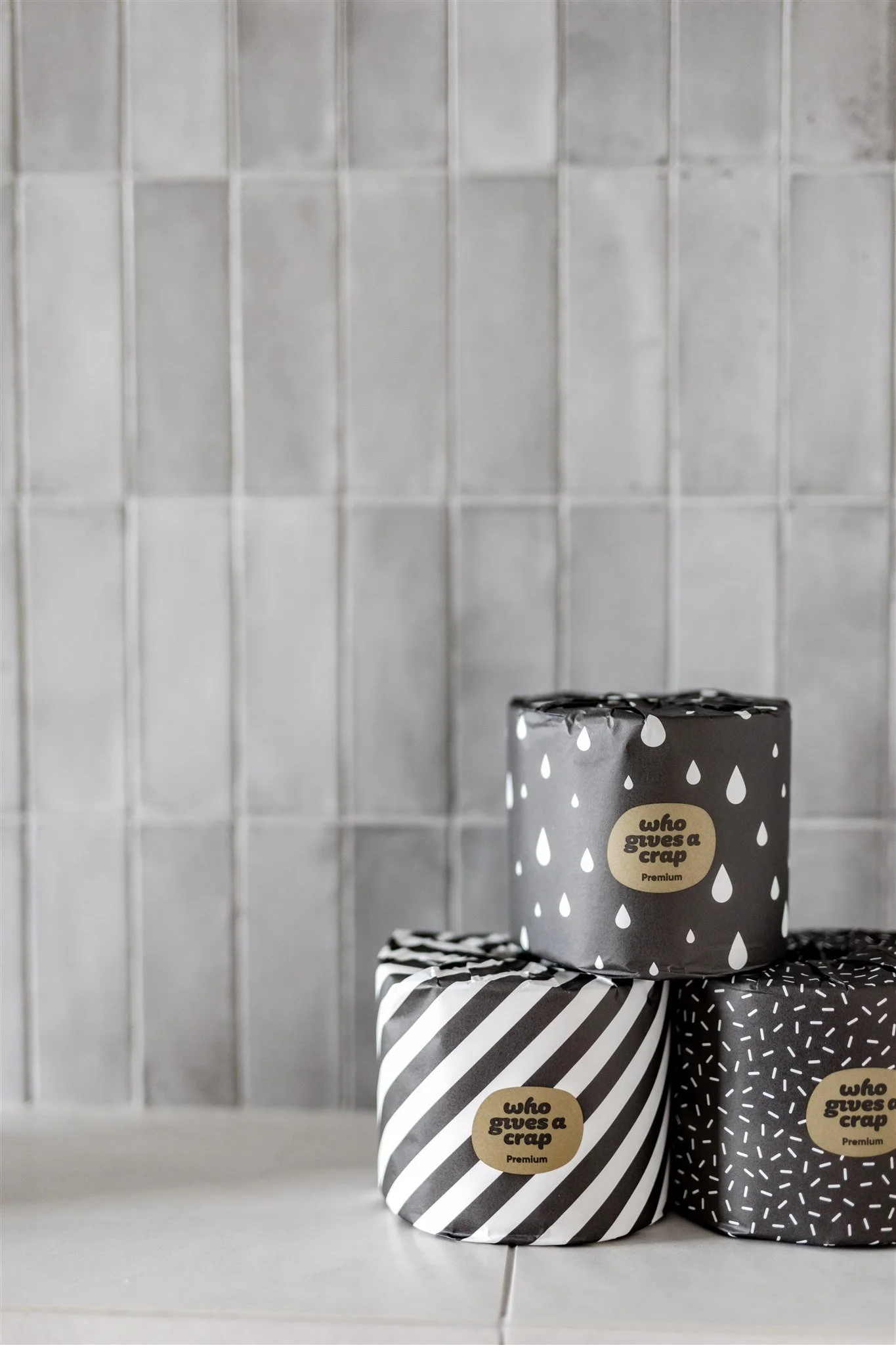 Three rolls of toilet paper with black and white patterned wrapping, placed on a tiled surface against a tiled wall, each roll has a beige label with black text that reads 'who gives a crap'.