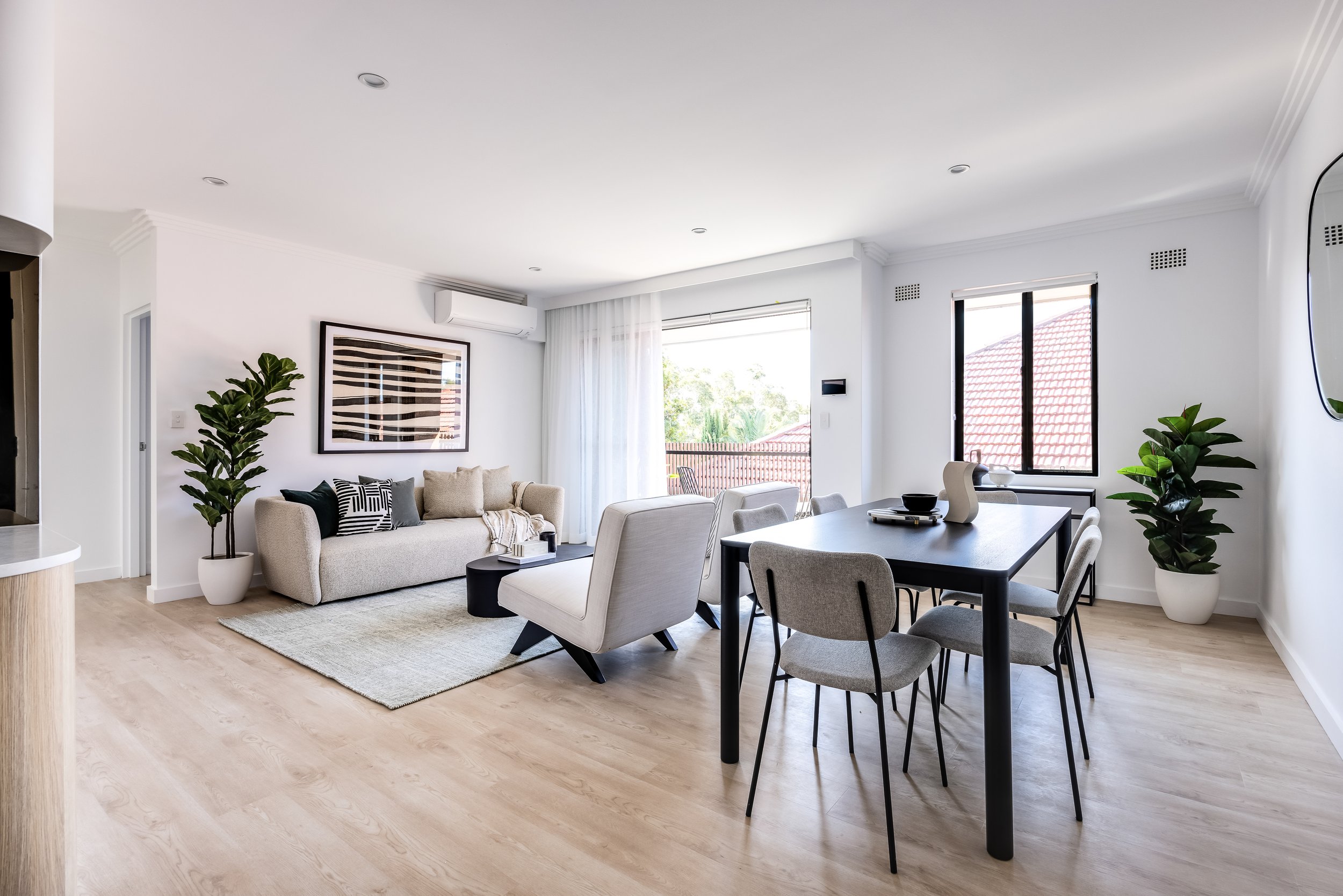 Modern living room with beige sofa and armchair, black coffee table, dining table with six gray chairs, large windows with white curtains, potted plants, and artwork on white walls.