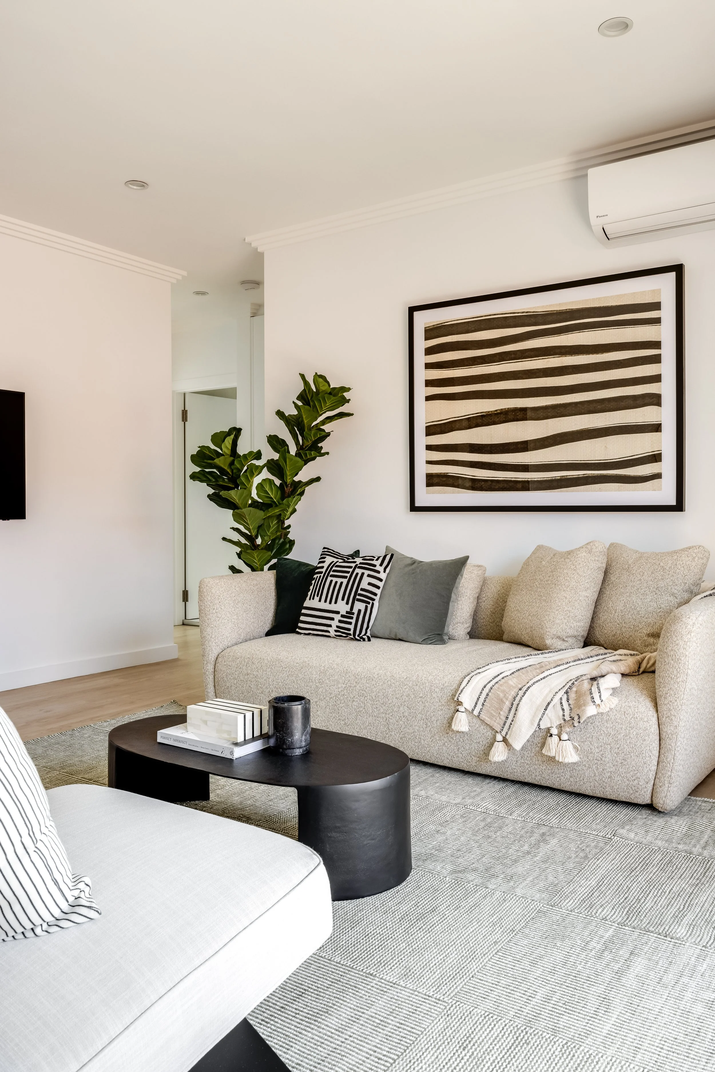 Modern living room with beige sofa, black coffee table, and decorative pillows, wall art with striped design, indoor plant, and air conditioning unit.