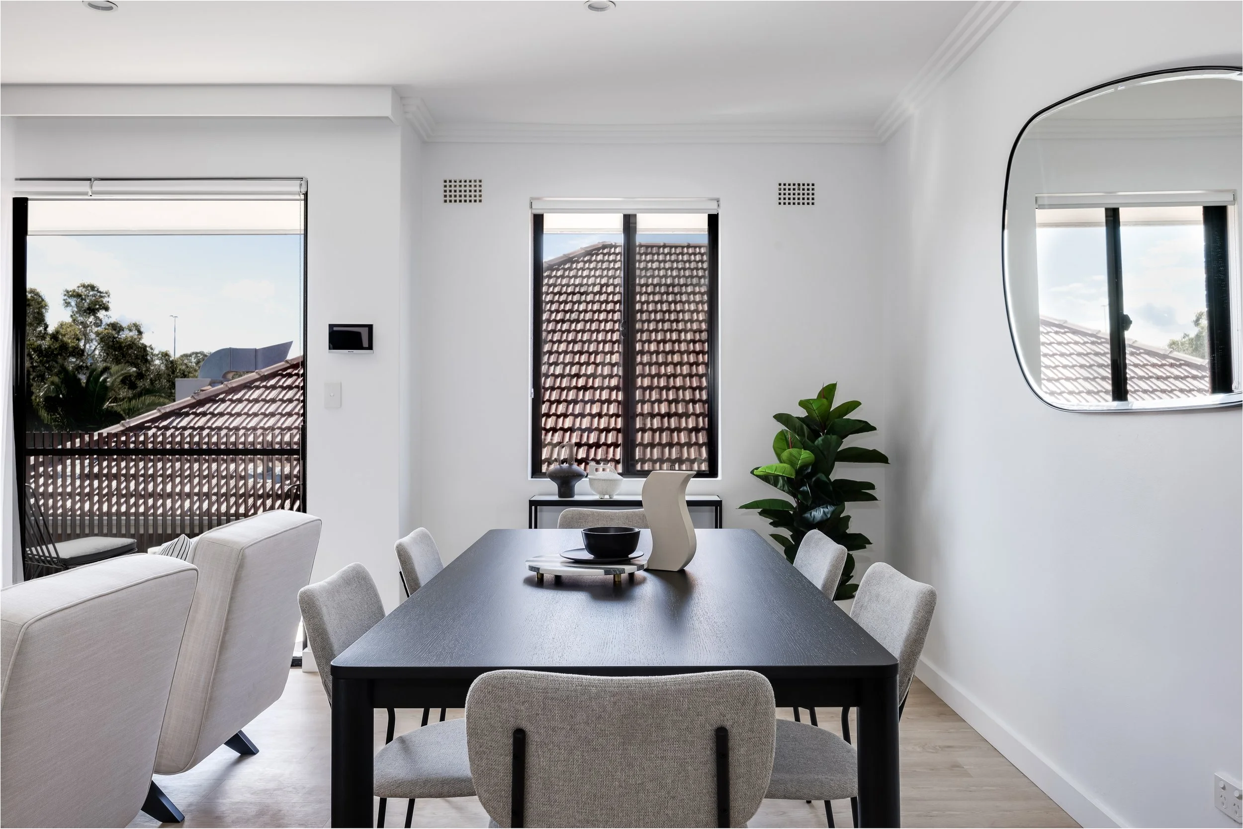 Modern dining room with black table, six light gray chairs, large windows showing rooftops, and a green plant in the corner.