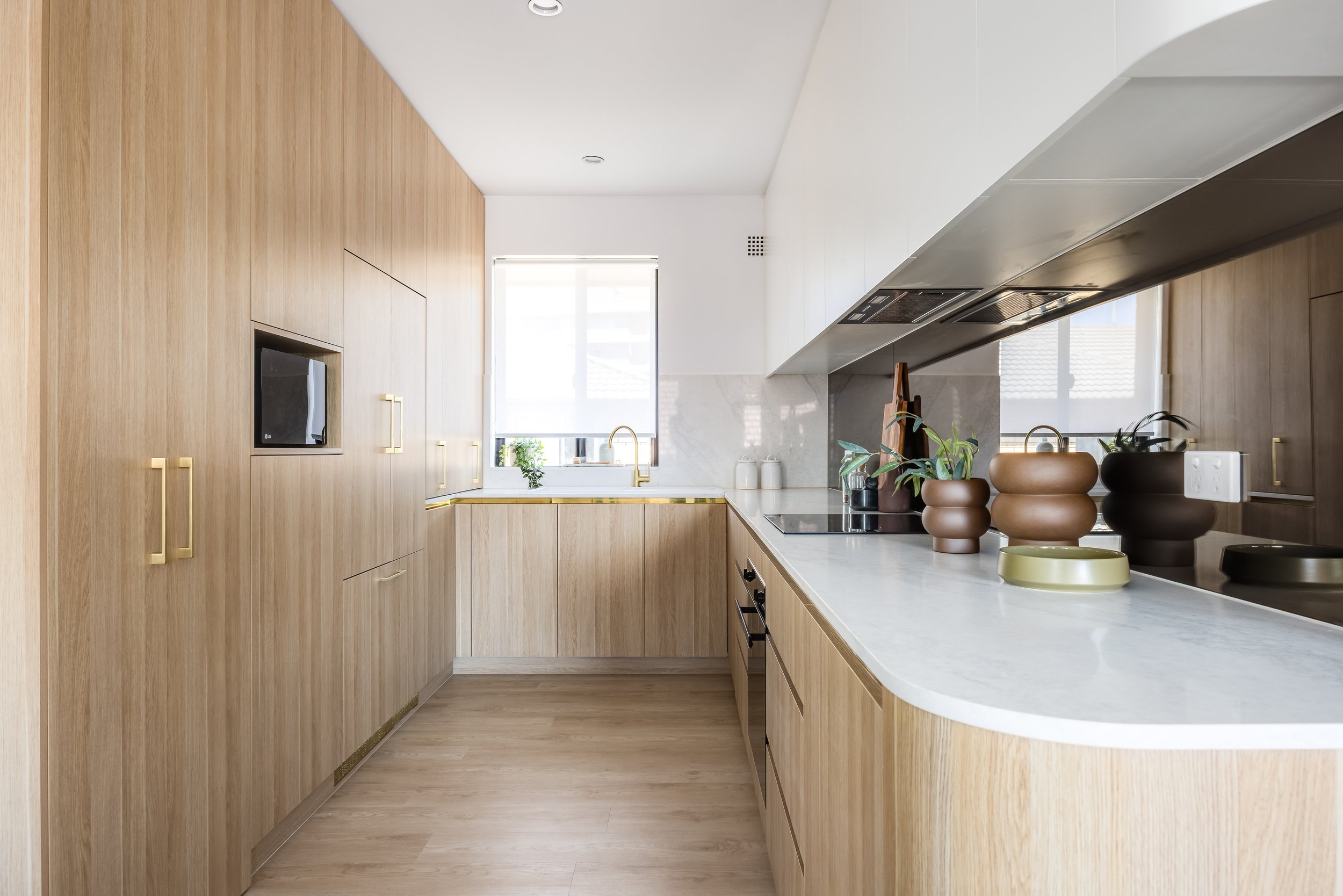 Modern kitchen with light wood cabinetry, white countertops, and gold hardware. There is a window with a small plant, and kitchen decor includes vases with plants.