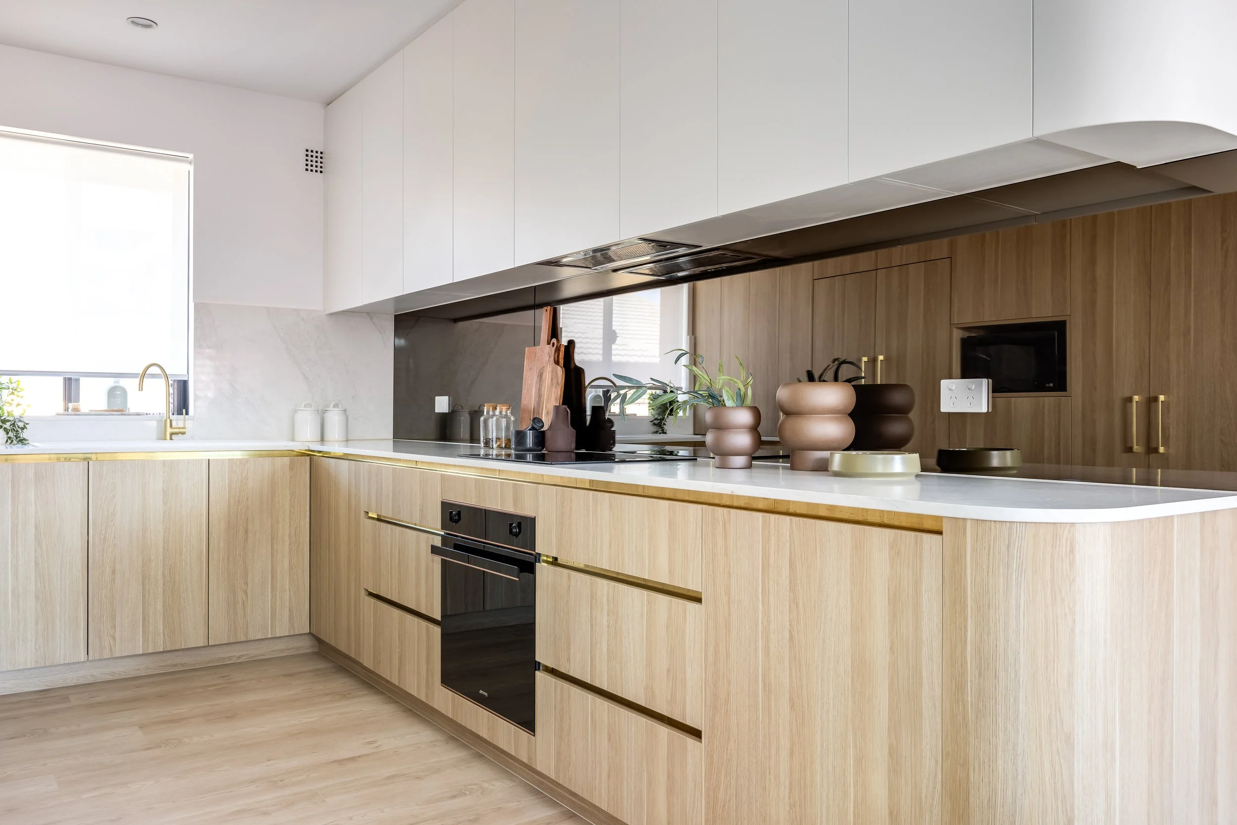 Modern kitchen with light wood cabinets, white countertops, and decorative vases; stainless steel oven; window with blinds; and a gold faucet.