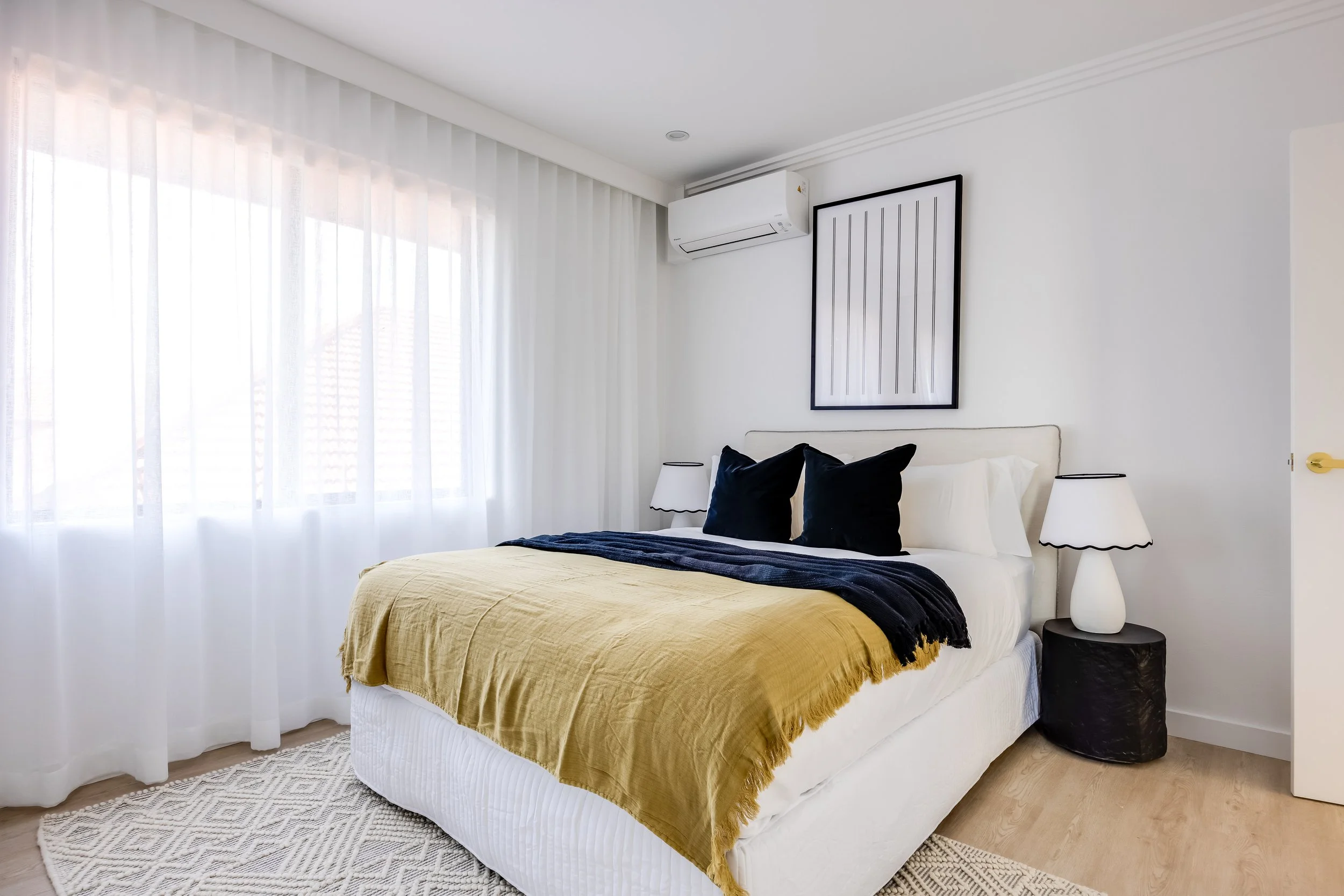 A modern bedroom with a large bed covered with a yellow blanket and black pillows, beside a black nightstand with a white lamp, white curtains, a framed artwork, and an air conditioning unit on the wall.
