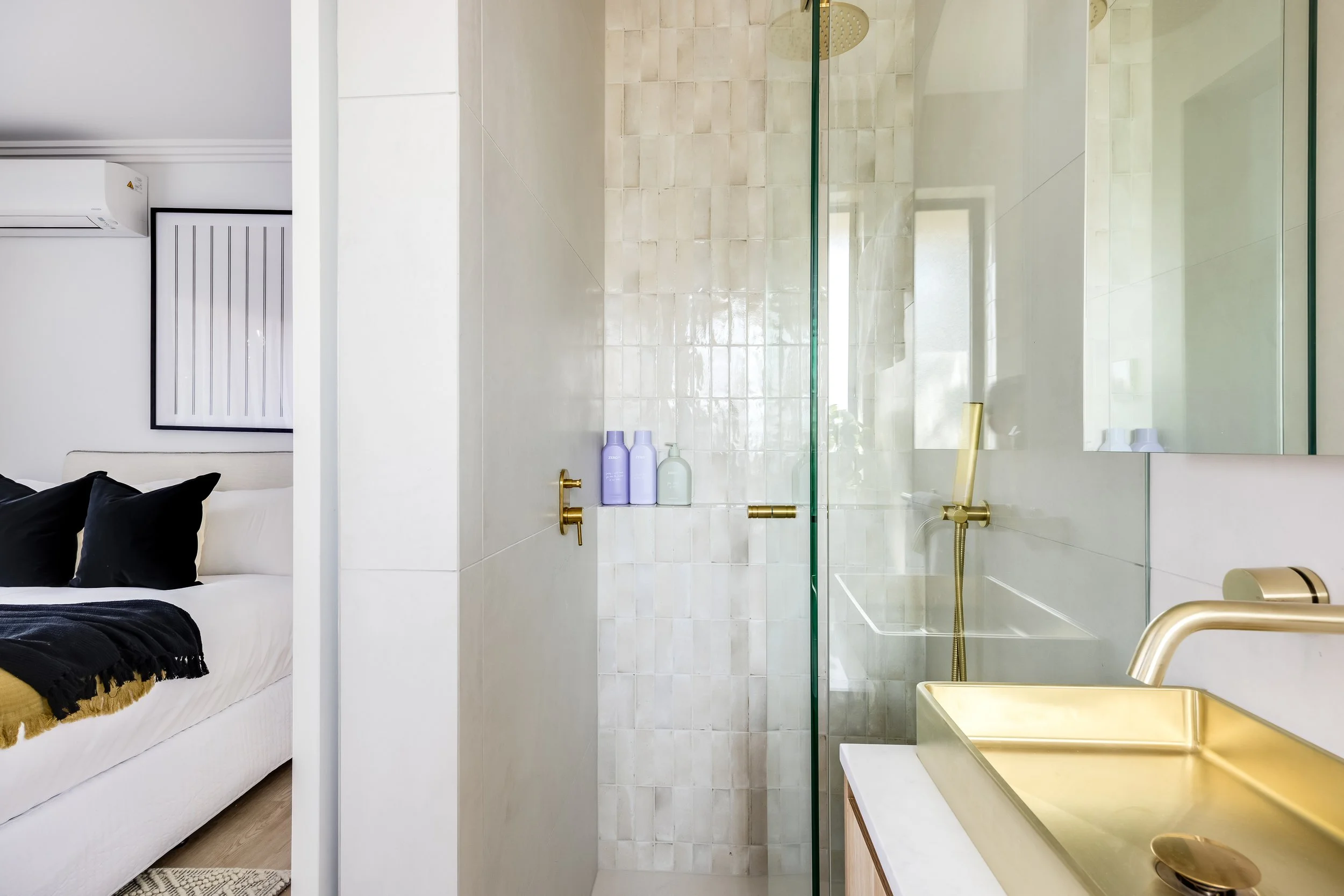 Interior of a modern bathroom with a gold sink and fixtures, glass shower enclosure, and bottles of toiletries, adjacent to a bedroom with a bed and black pillows.