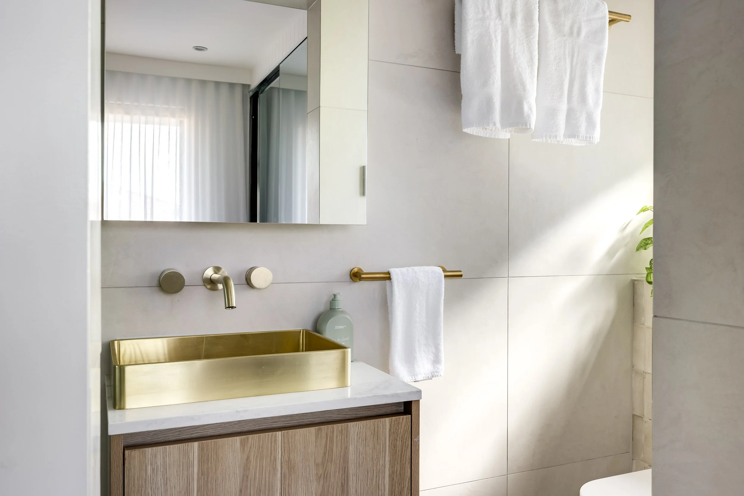 Modern bathroom with a gold sink, mirror, and white towels on a gold towel rack. Natural light coming through a window with white curtains, and a small plant on the right