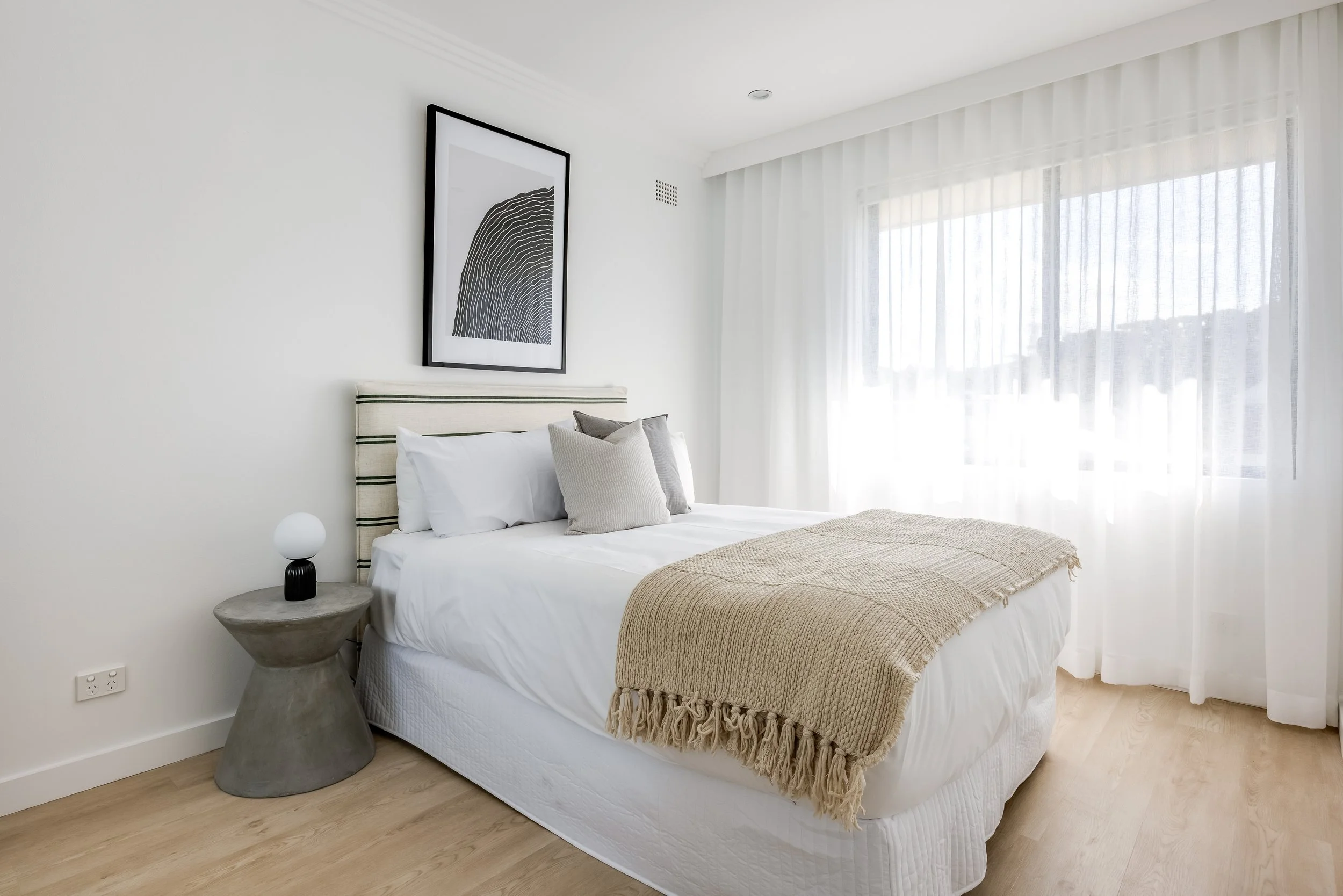 Bright modern bedroom with a white bed, beige blanket, decorative pillows, a concrete side table with a lamp, minimalist black and white artwork, and large window with sheer white curtains.