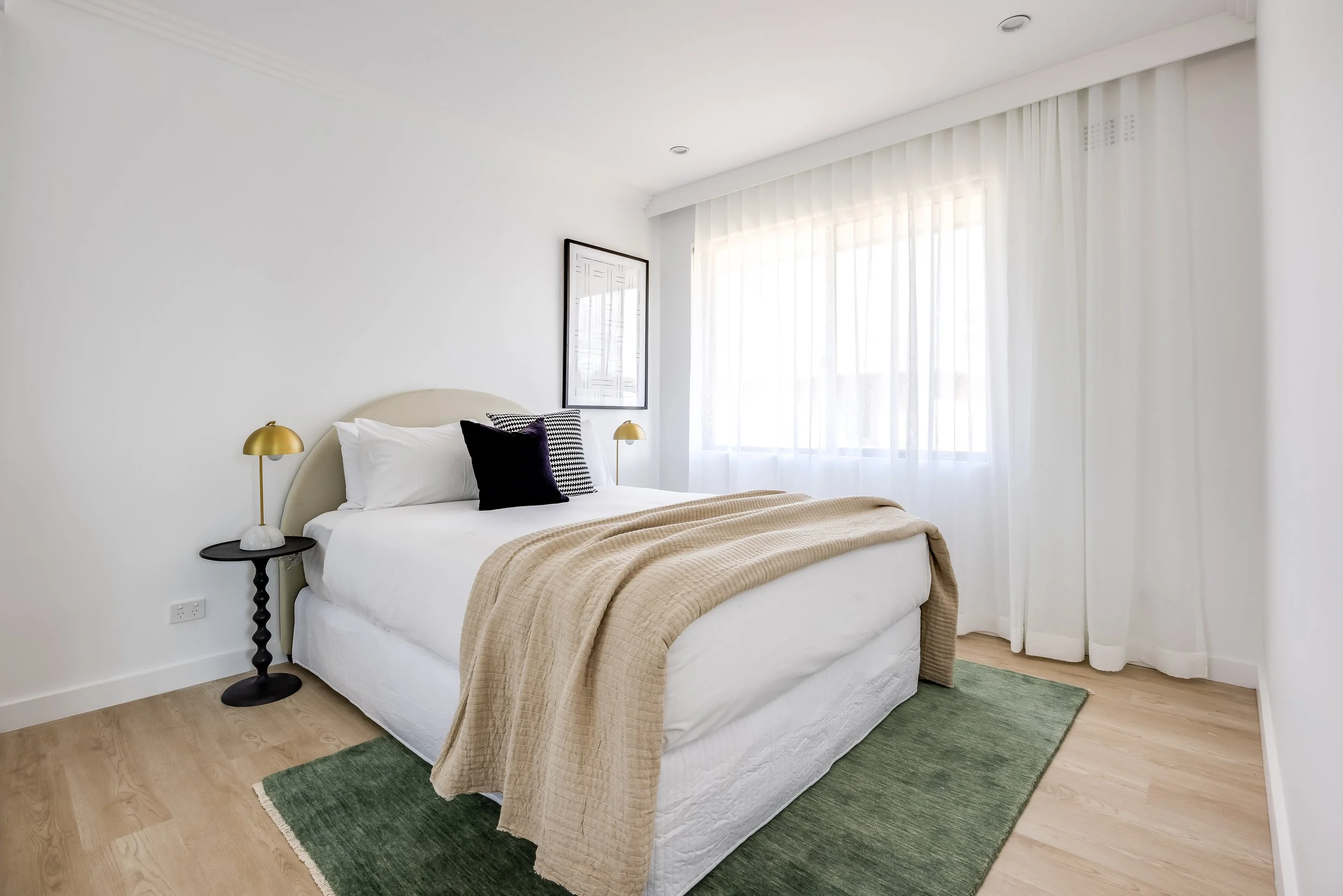 A bedroom with a white bed, black and gold bedside lamps, dark framed wall art, white curtains, a beige blanket, black pillow, and a green rug.
