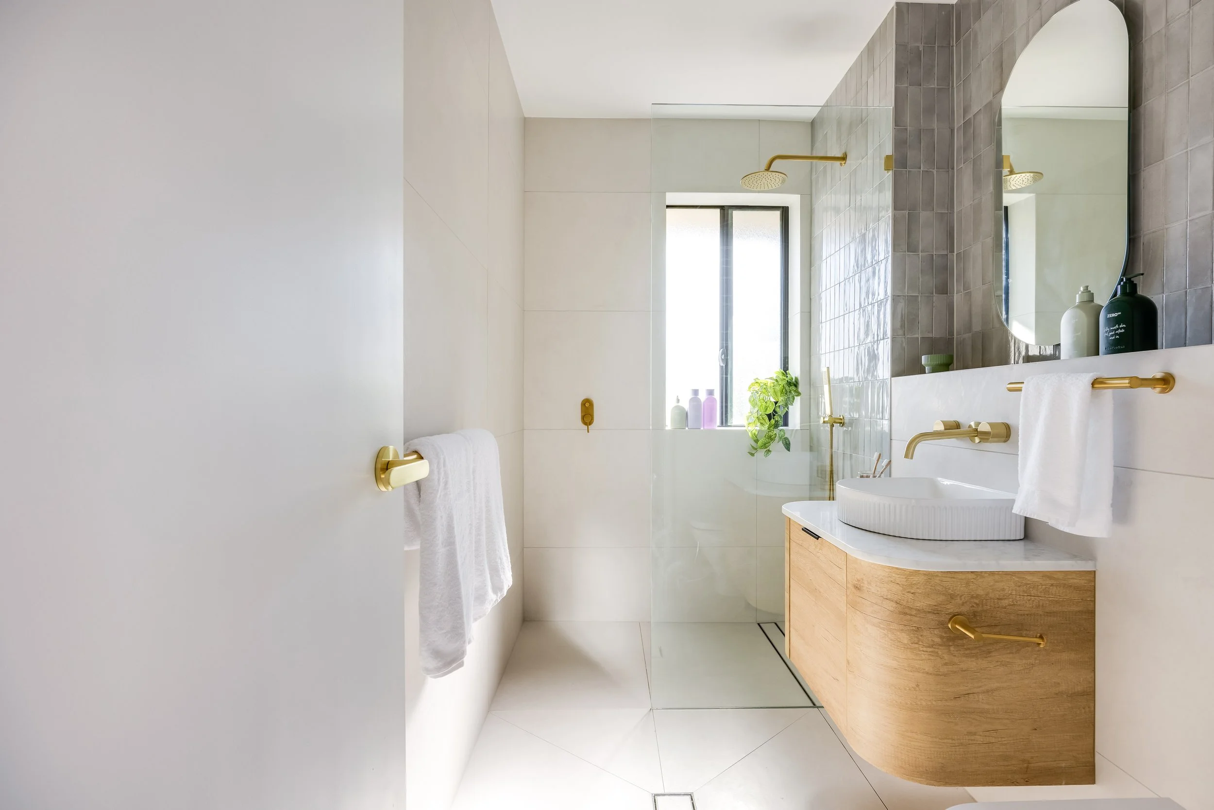 Modern bathroom with white walls, wooden vanity, round white sink, gold fixtures, large mirror, glass shower with gold showerhead, and a window with plant and bottles.