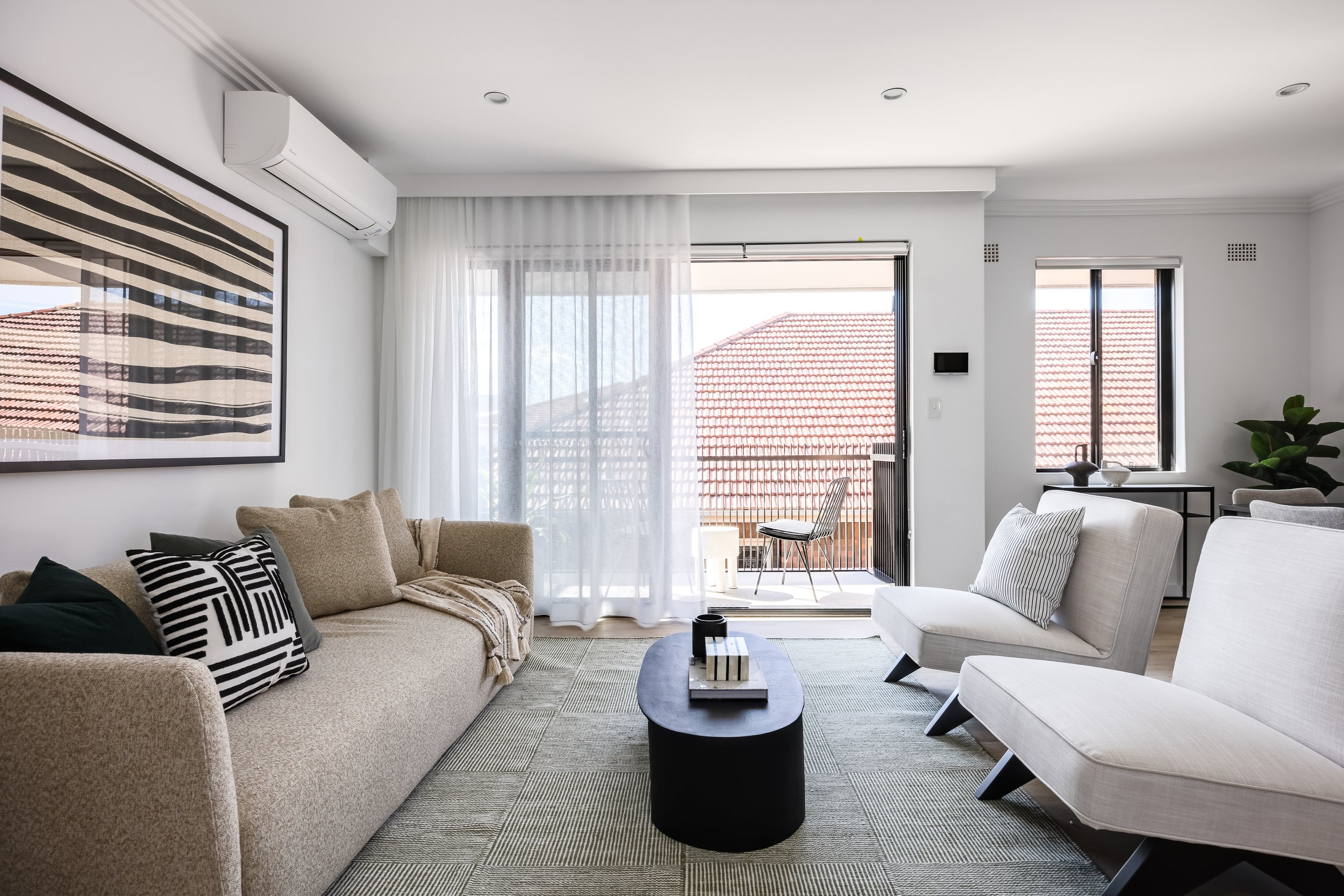Modern living room with a beige couch, white armchairs, black and white pillows, a black coffee table, and a balcony with outdoor chairs and a red-tiled roof visible through large glass sliding doors.