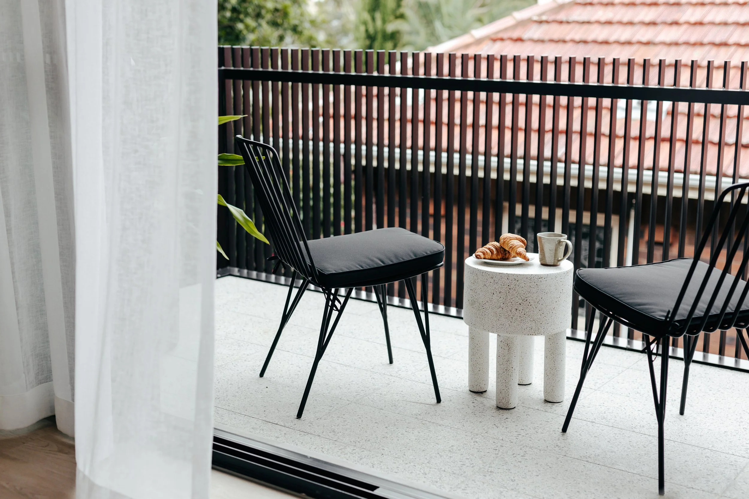 Balcony porch with two black chairs, a small white table holding croissants and a mug, view of red roof tiles and greenery outside, white curtain on the left.