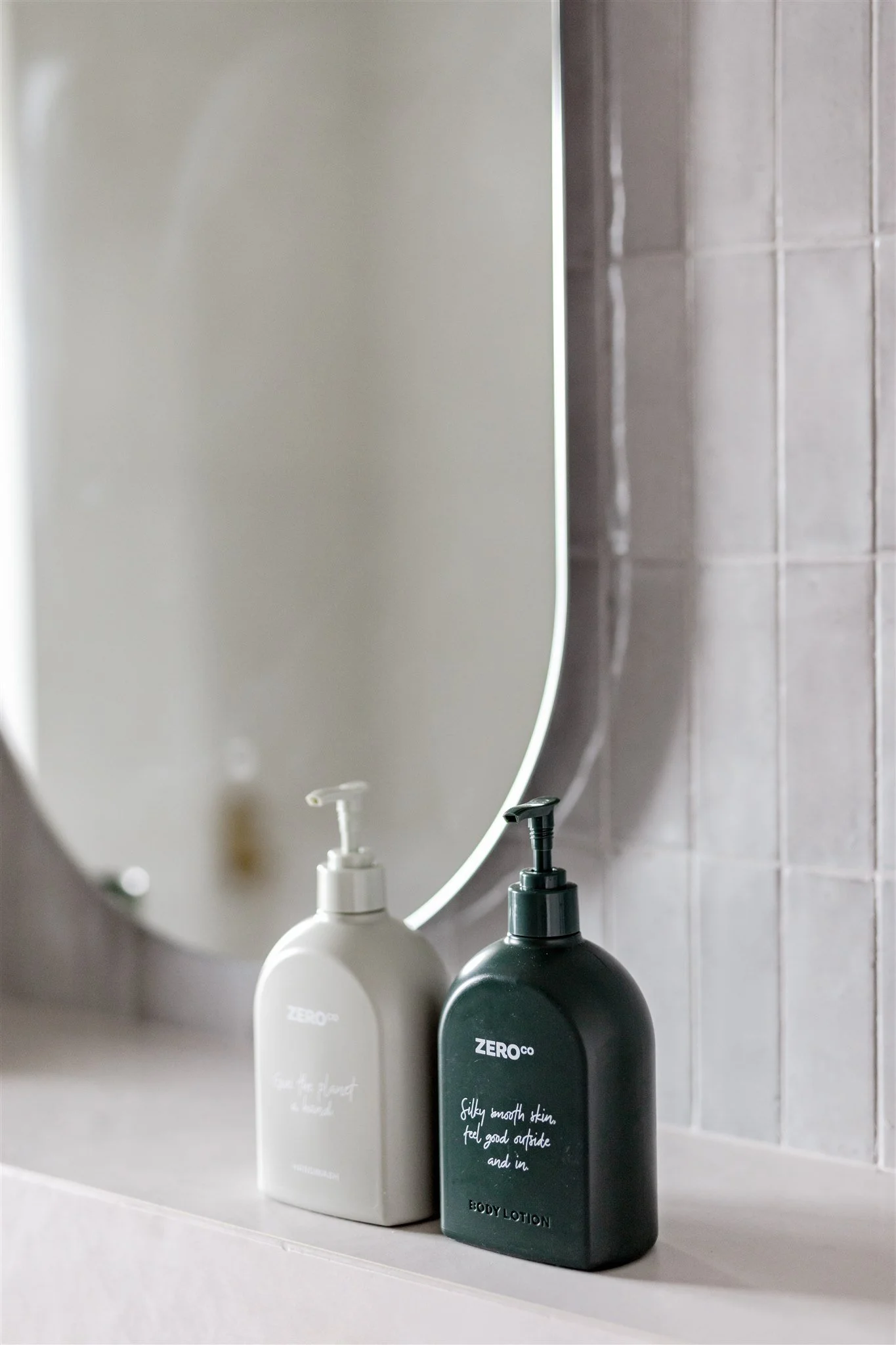 Two bottles of lotion, one white and one black, placed on a bathroom shelf in front of an oval mirror and tiled wall.