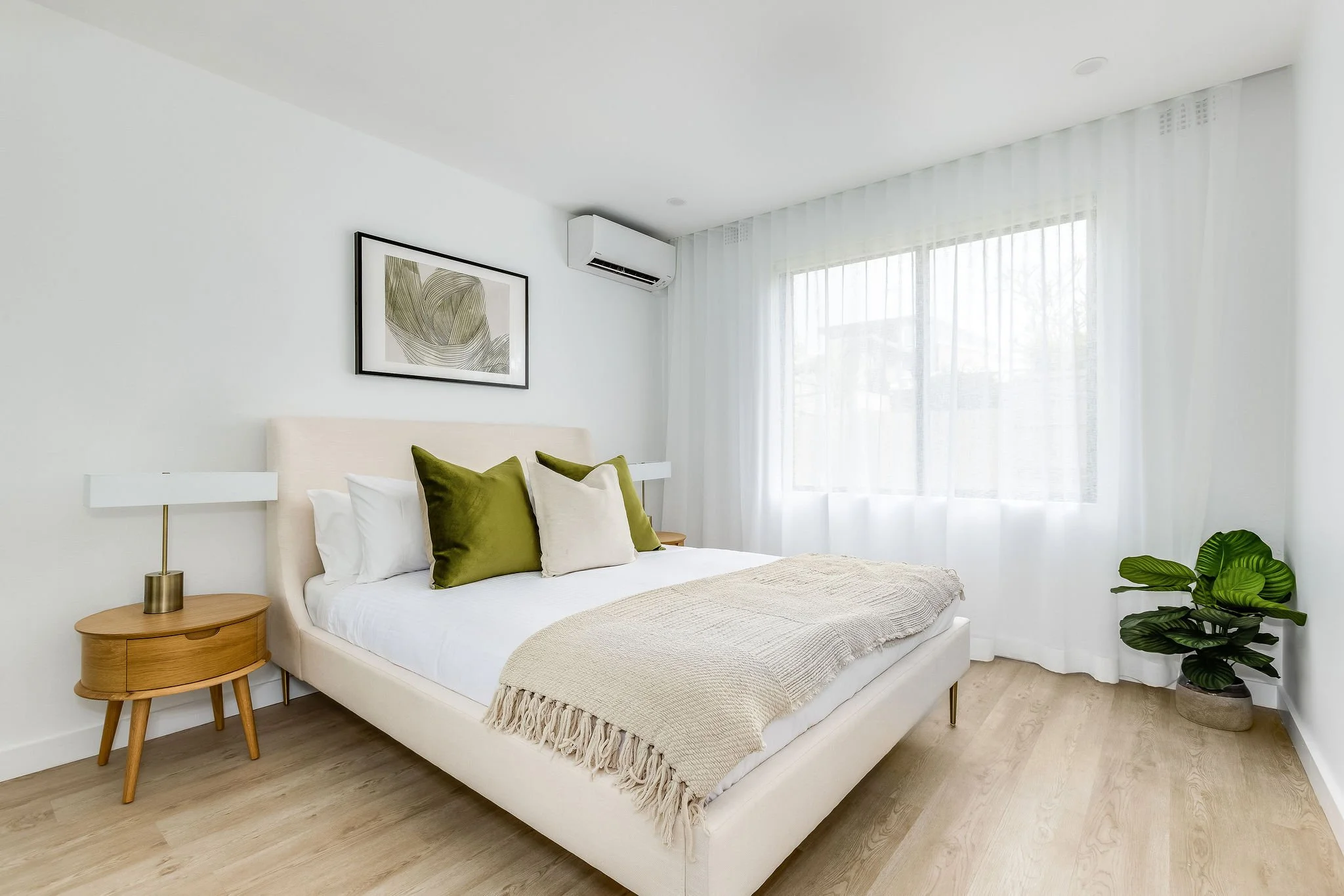 A minimalist bedroom with a white bed, green and beige pillows, a beige throw blanket, wooden nightstands, white lamps, a framed botanical artwork, a window with sheer white curtains, and a potted green plant.