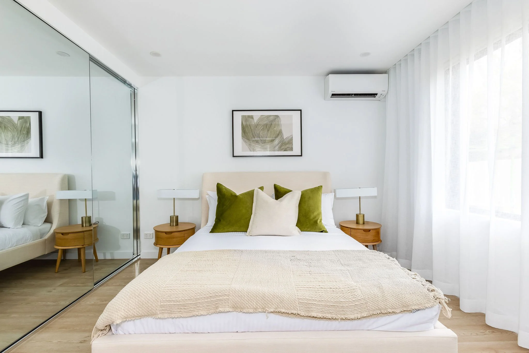 A neatly made bed with green and beige pillows in a modern bedroom with white walls, wooden bedside tables with lamps, and a large window with white curtains.