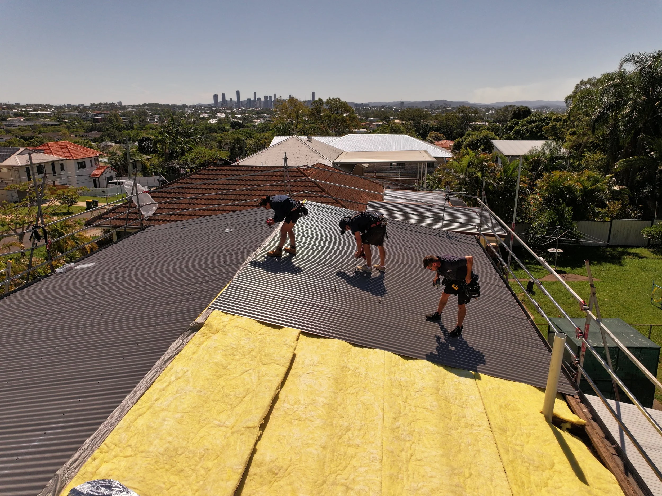 Workers installing roofing material on a house with a city skyline in the background.