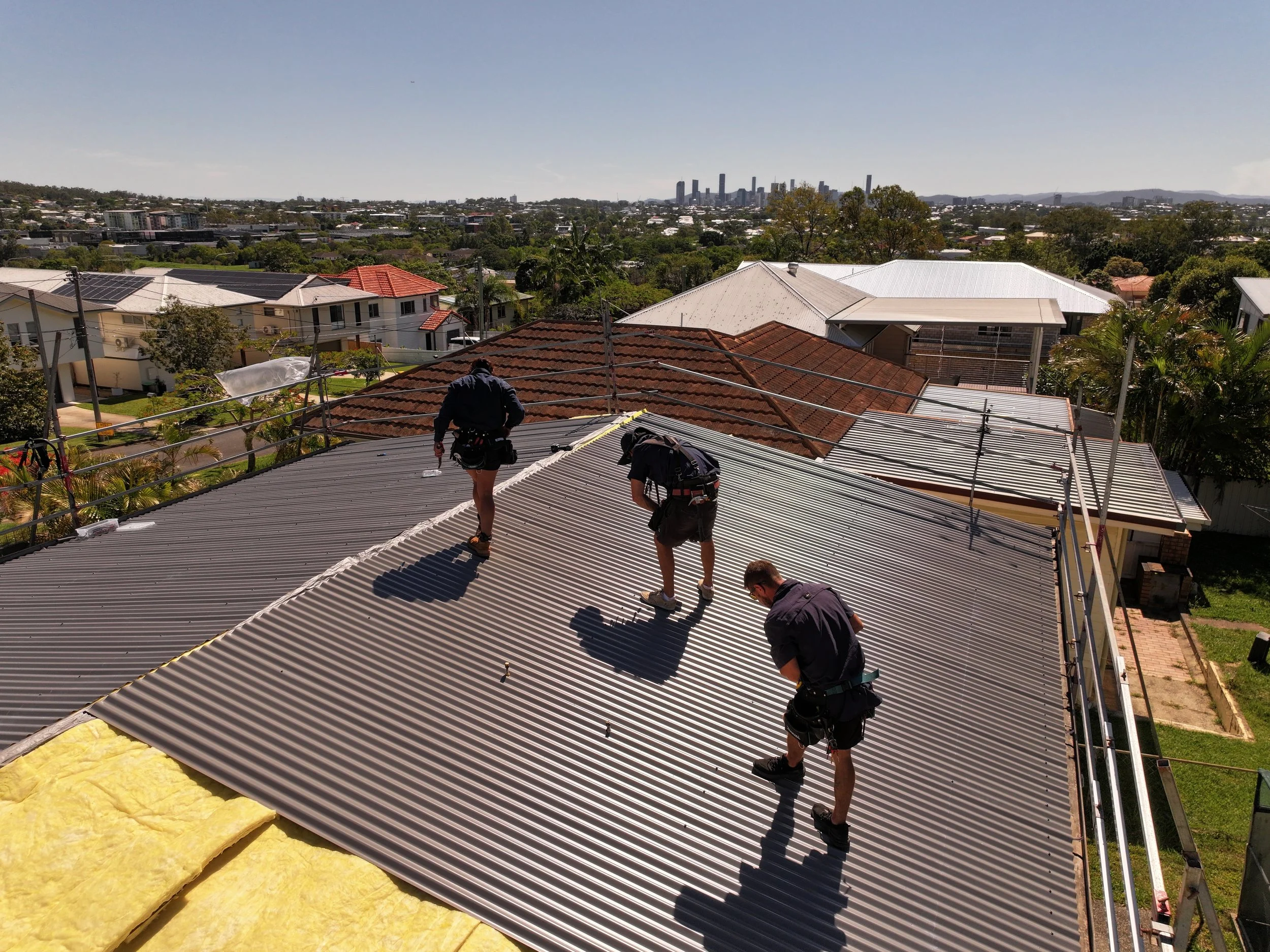 Workers installing corrugated metal roofing on a residential building with a view of a city skyline in the distance.