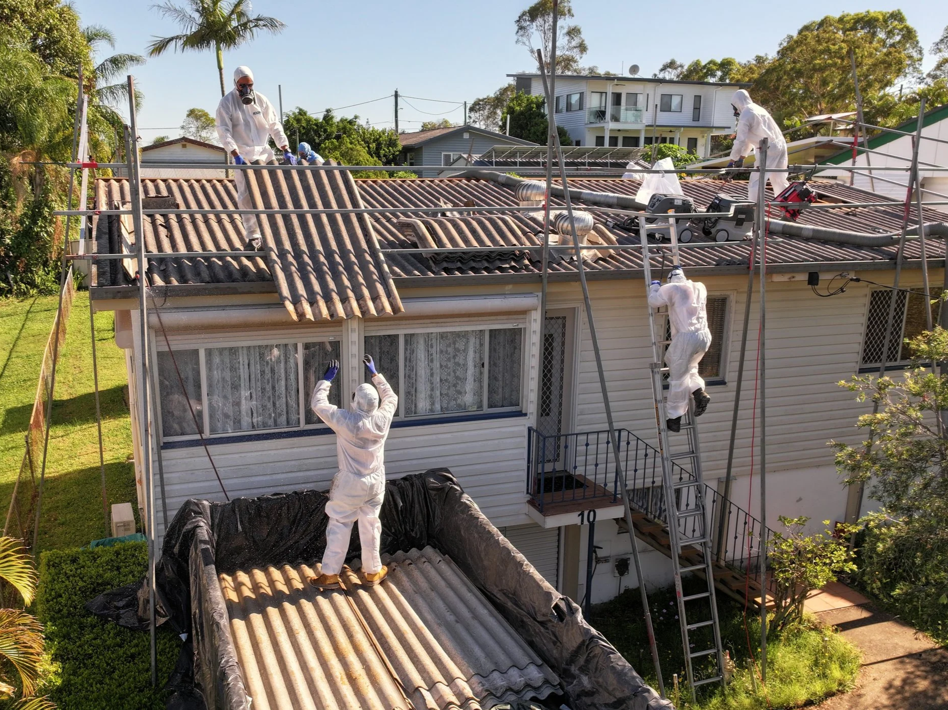 Workers in protective suits and masks repairing or inspecting a house roof with scaffolding and ventilation systems.