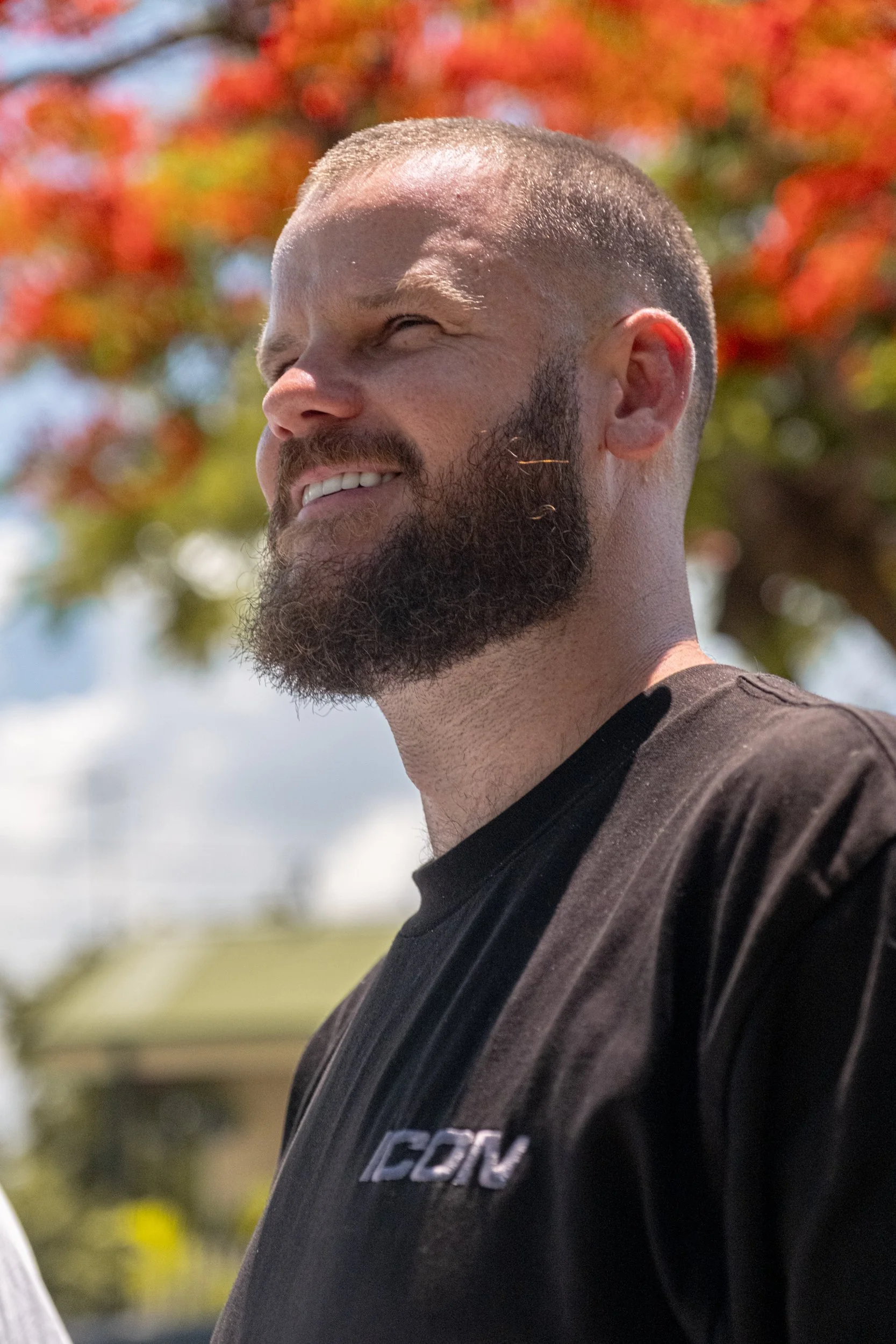 A man with a beard and short hair smiling outdoors, with a background of colorful autumn leaves.