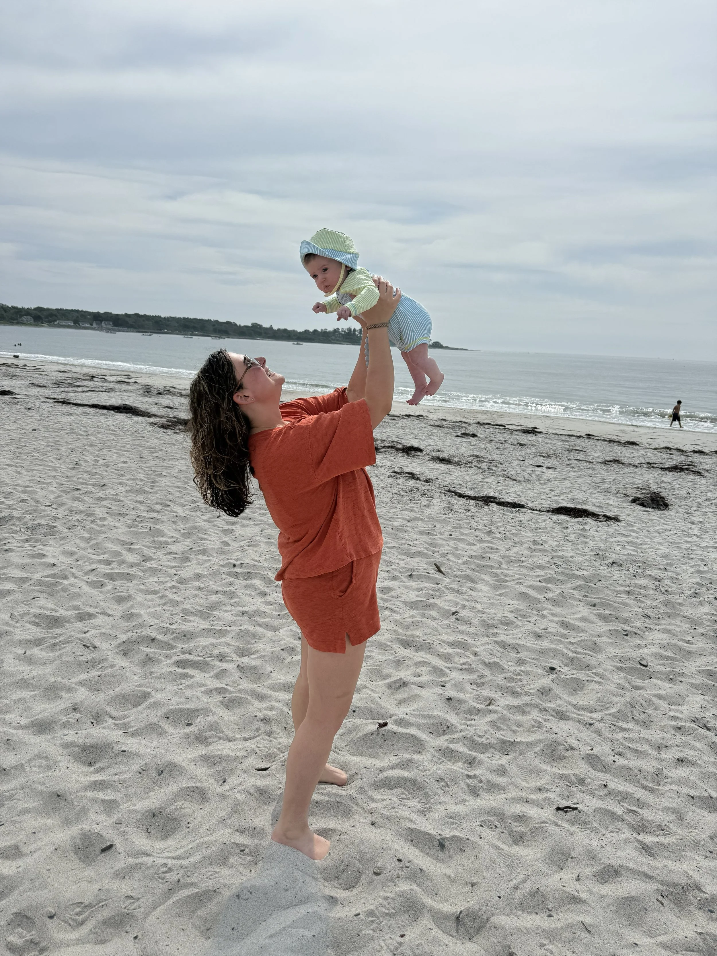 A woman in an orange outfit playing with a baby on the beach, lifting the baby into the air with the ocean and cloudy sky in the background.