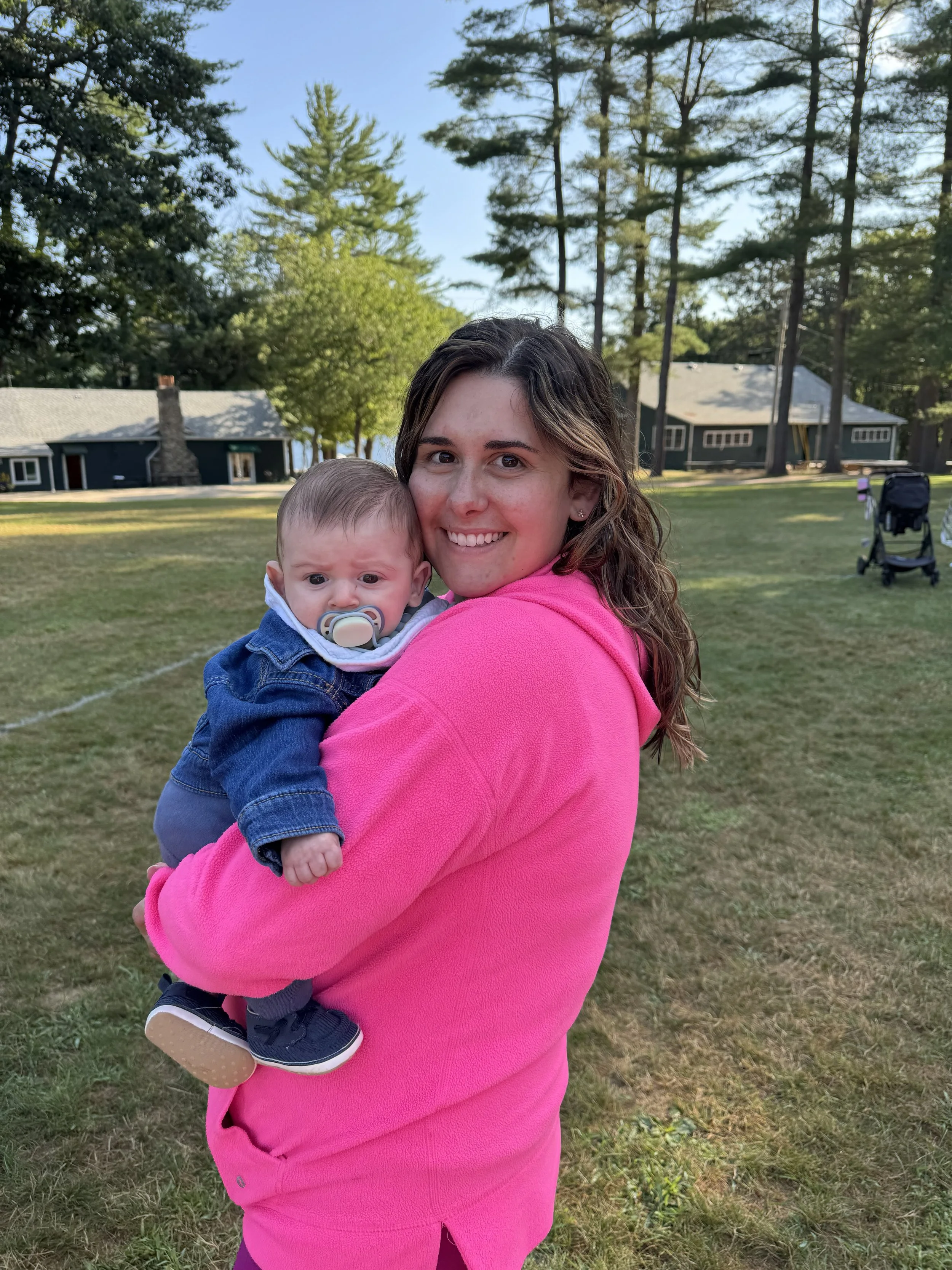 A young woman with long wavy hair wearing a pink hoodie, holding a baby with a pacifier, outdoors in a grassy area with trees and houses in the background.