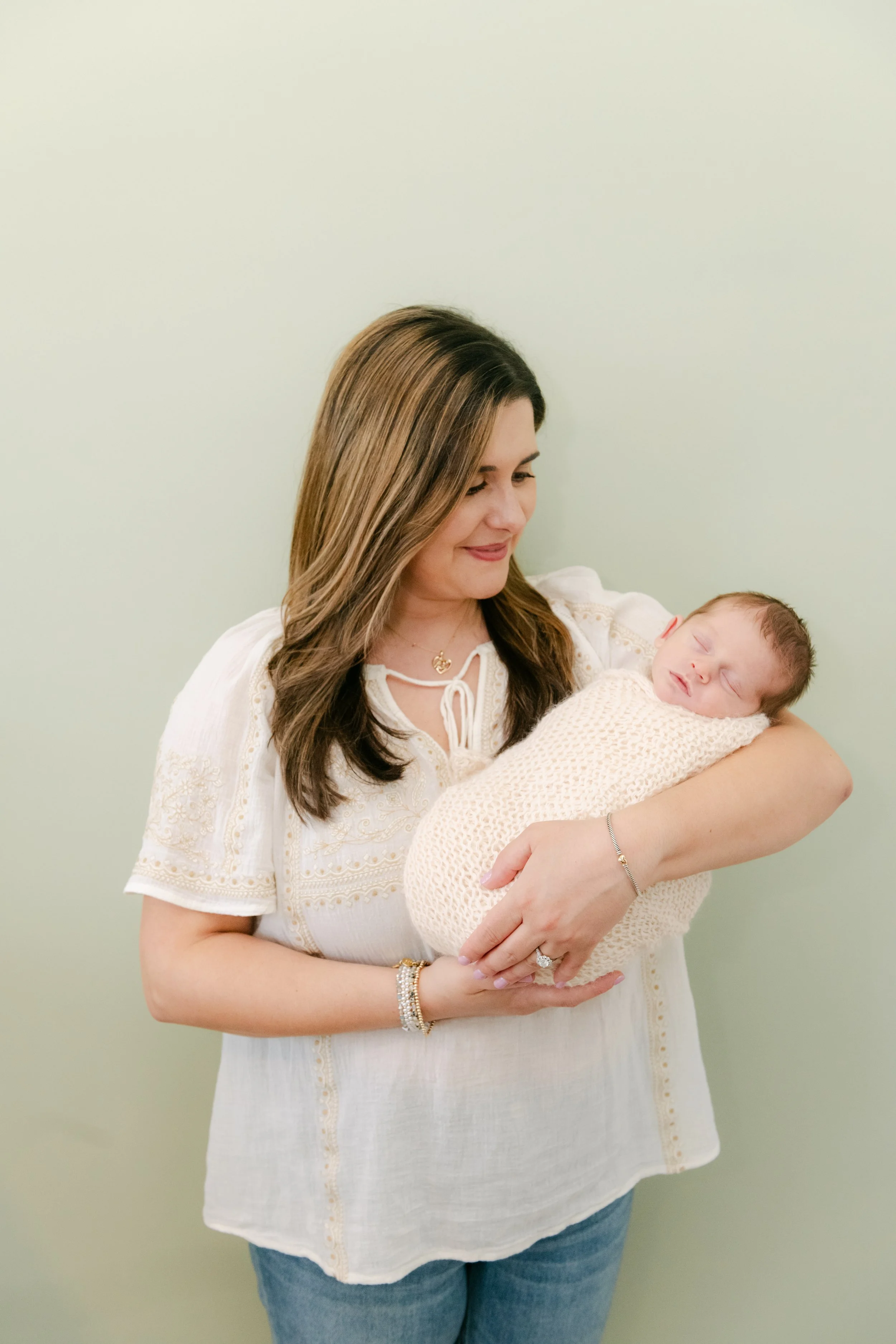 Woman holding a sleeping baby wrapped in a cozy blanket, standing against a light green wall.