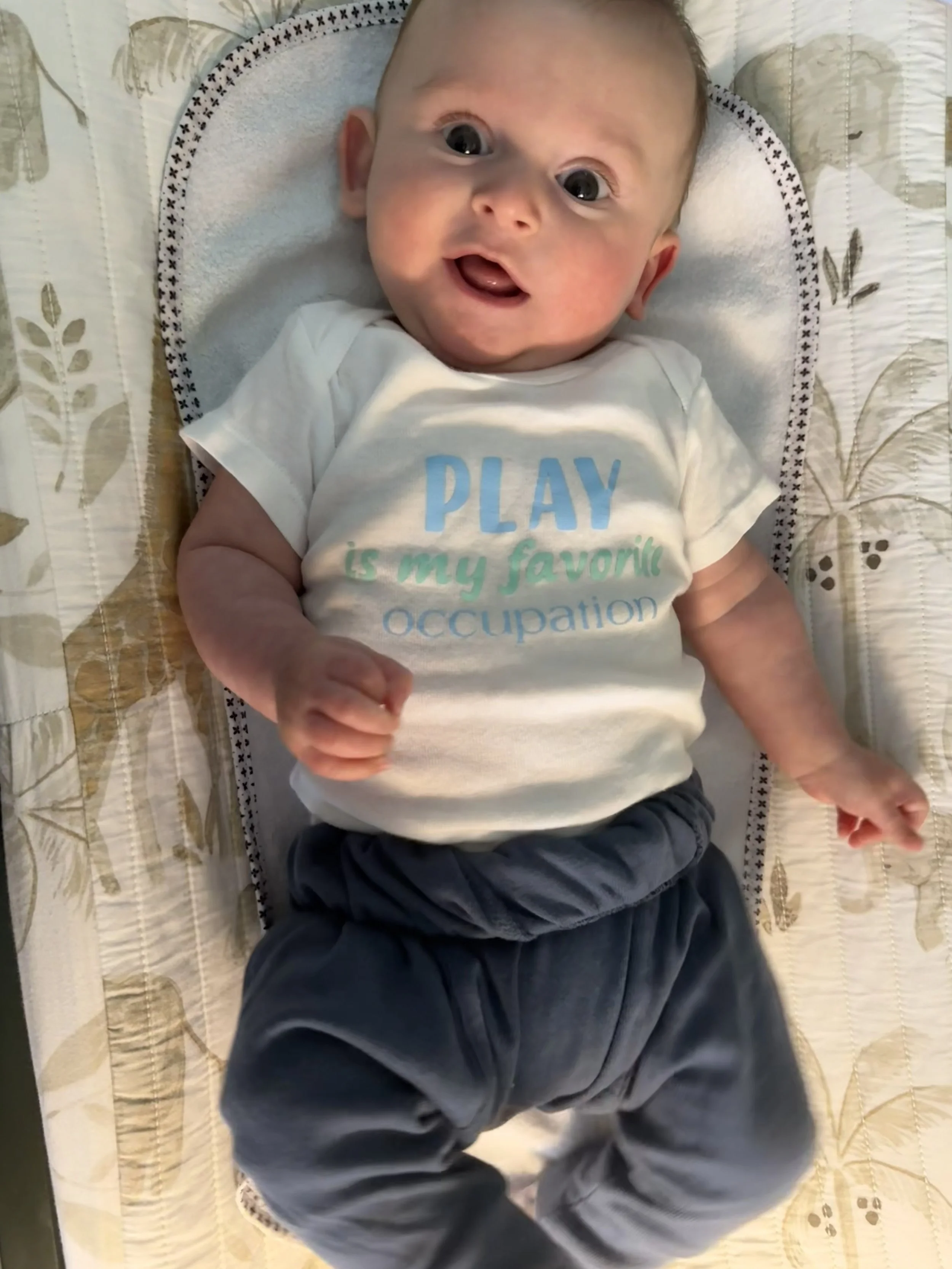 A baby lying on a quilted mattress, wearing a white t-shirt with a phrase about play and dark pants, with a happy expression and mouth open.